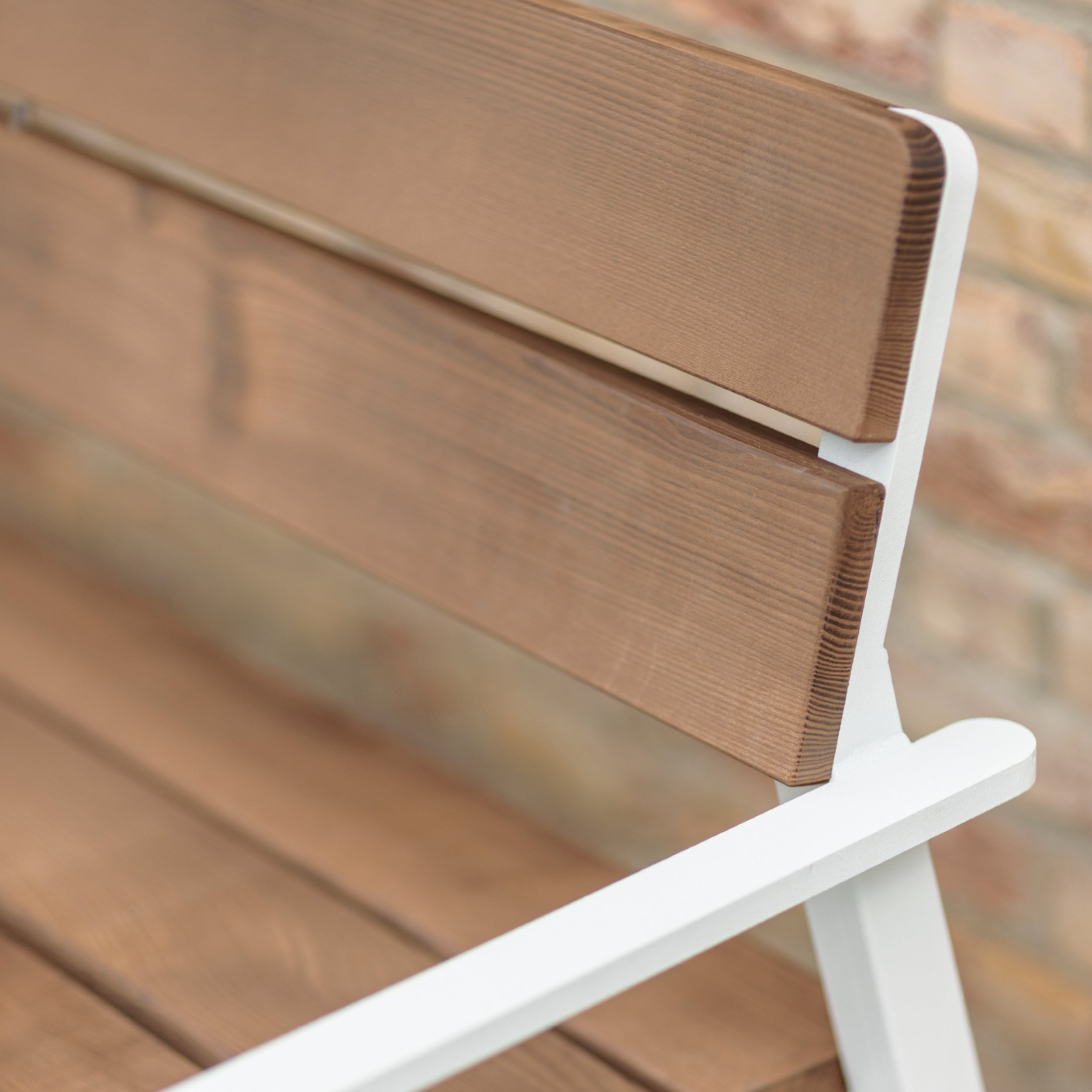 Close-up of an Egoé Cora Public Bench showcasing Jatoba wood slats and a sleek white metal frame, set against a blurred brick wall. The natural wood finish complements the white metal, creating a striking contrast.