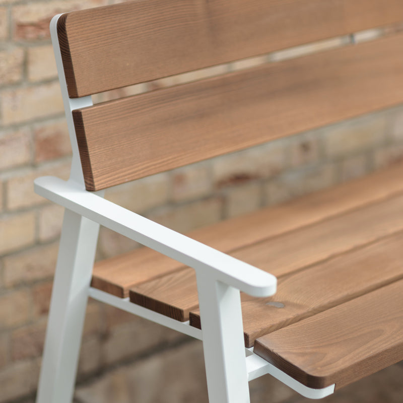 Close-up of the Cora Public Chair by Egoe, showcasing a simple modern design with smooth brown Jatoba wood slats and a white metal frame with corrosion-protective zinc. Its positioned against a light brick wall.