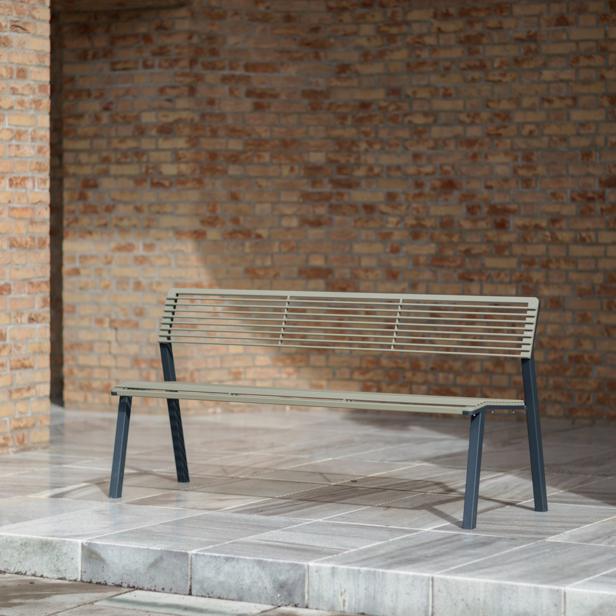 The Cora Public Bench, by Egoé, is a minimalist metal piece crafted from powder-coated steel. It stands on tiles against a brick wall, showcasing its slatted design that embodies the simplicity and modernity typical of Egoé street furniture.