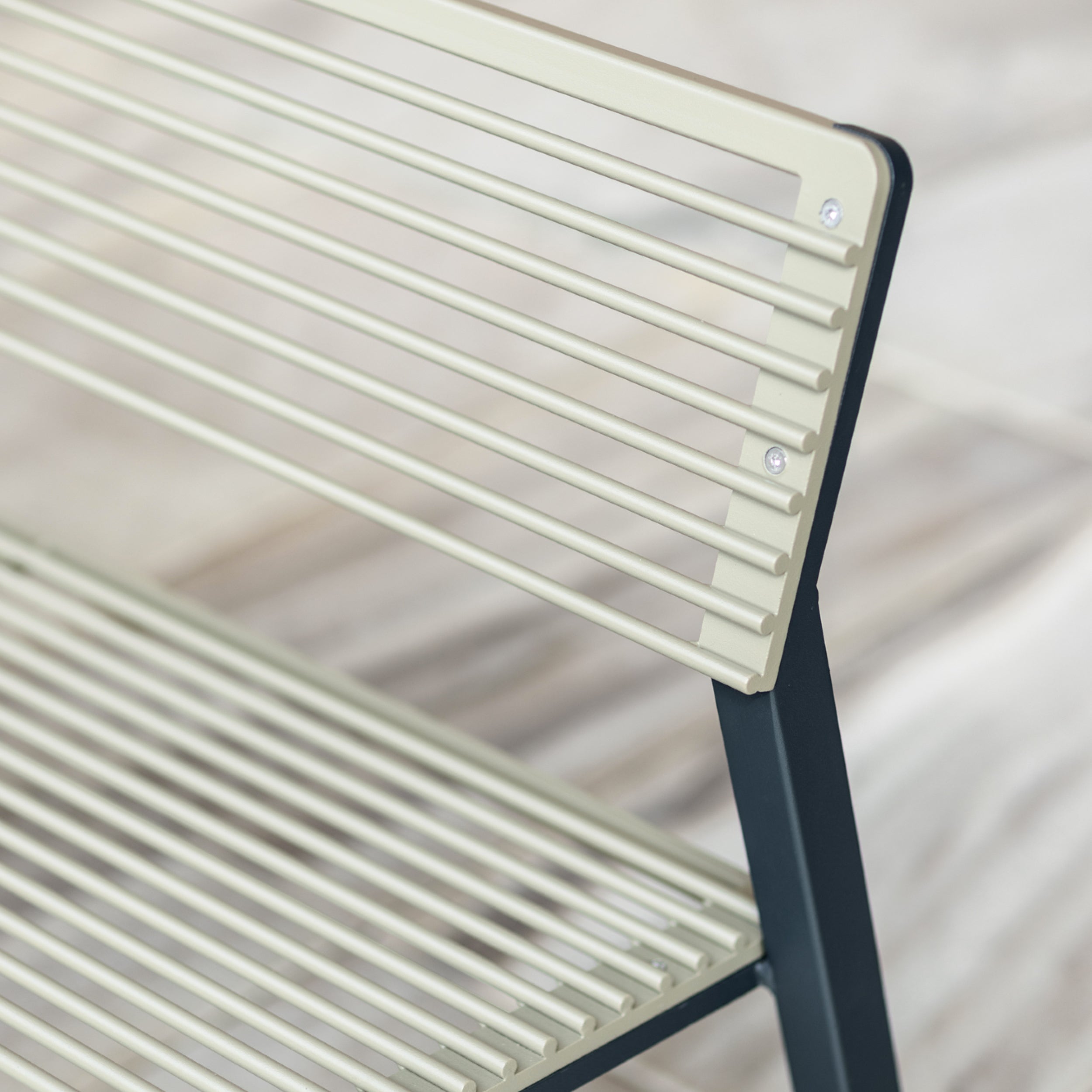A close-up of the Cora Public Bench by Egoe shows a modern design with a light beige slatted backrest and seat made from tropical Jatoba wood, supported by a sleek black frame against a blurred, neutral-toned background, evoking minimalist elegance.