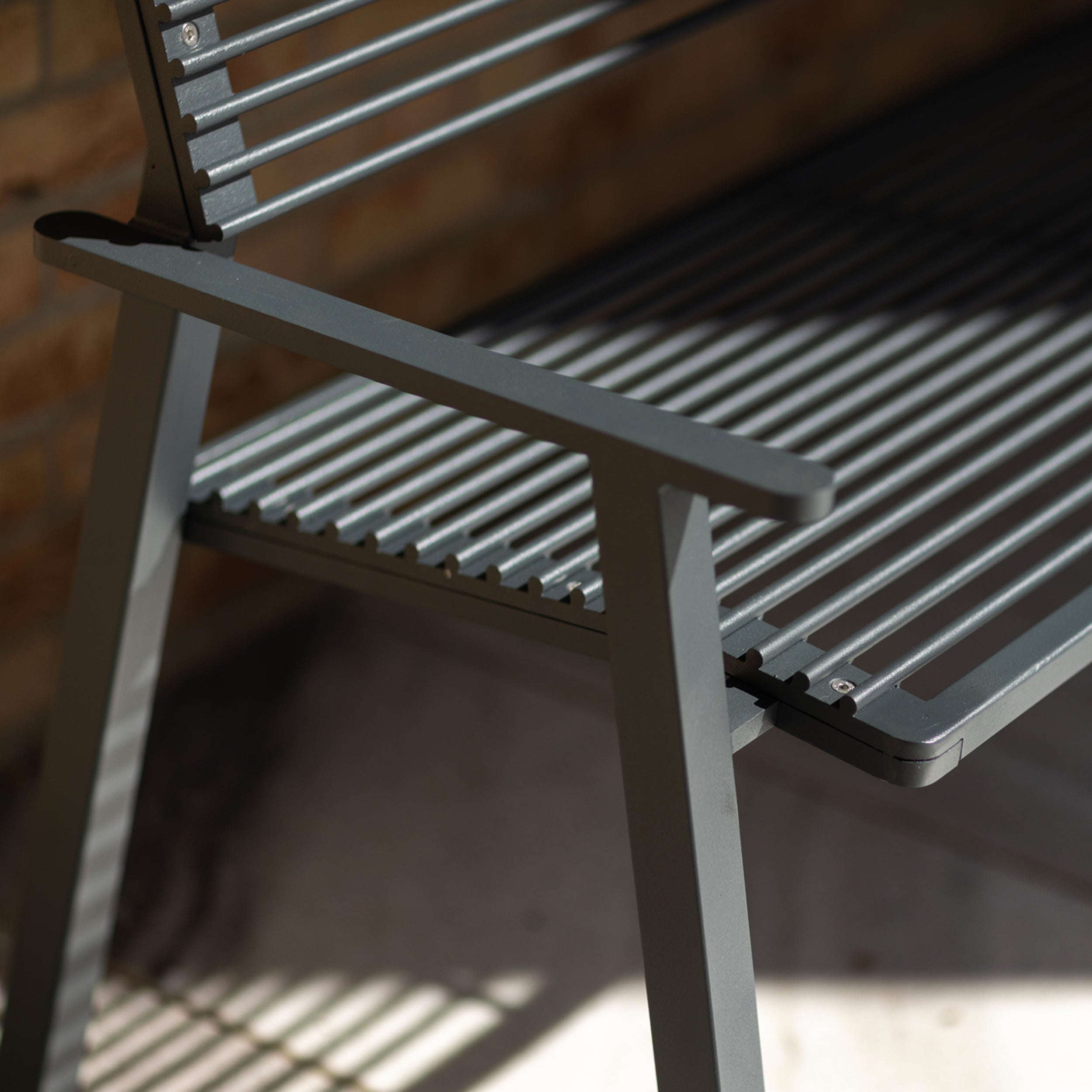 A close-up of the Egoe Cora Public Bench, crafted from zinc and powder-coated steel with slatted seat and backrest, shows sunlight casting narrow shadows across its surface, creating a striped pattern. The bench is set near a brick wall on a patio or deck.