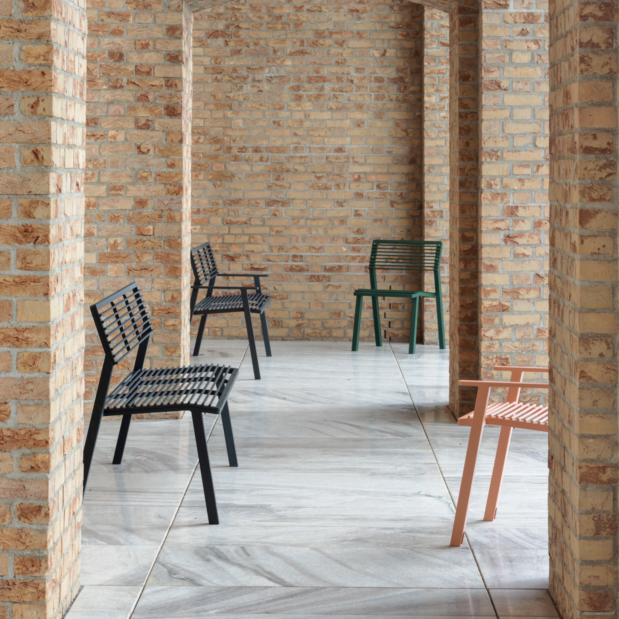 A brick-walled corridor showcases four colorful modern chairs with minimalist designs. These Cora Public Benches by Egoé, crafted from zinc and powder-coated steel, come in black, green, and peach, beautifully contrasting with the rustic brick arches and marble floor.