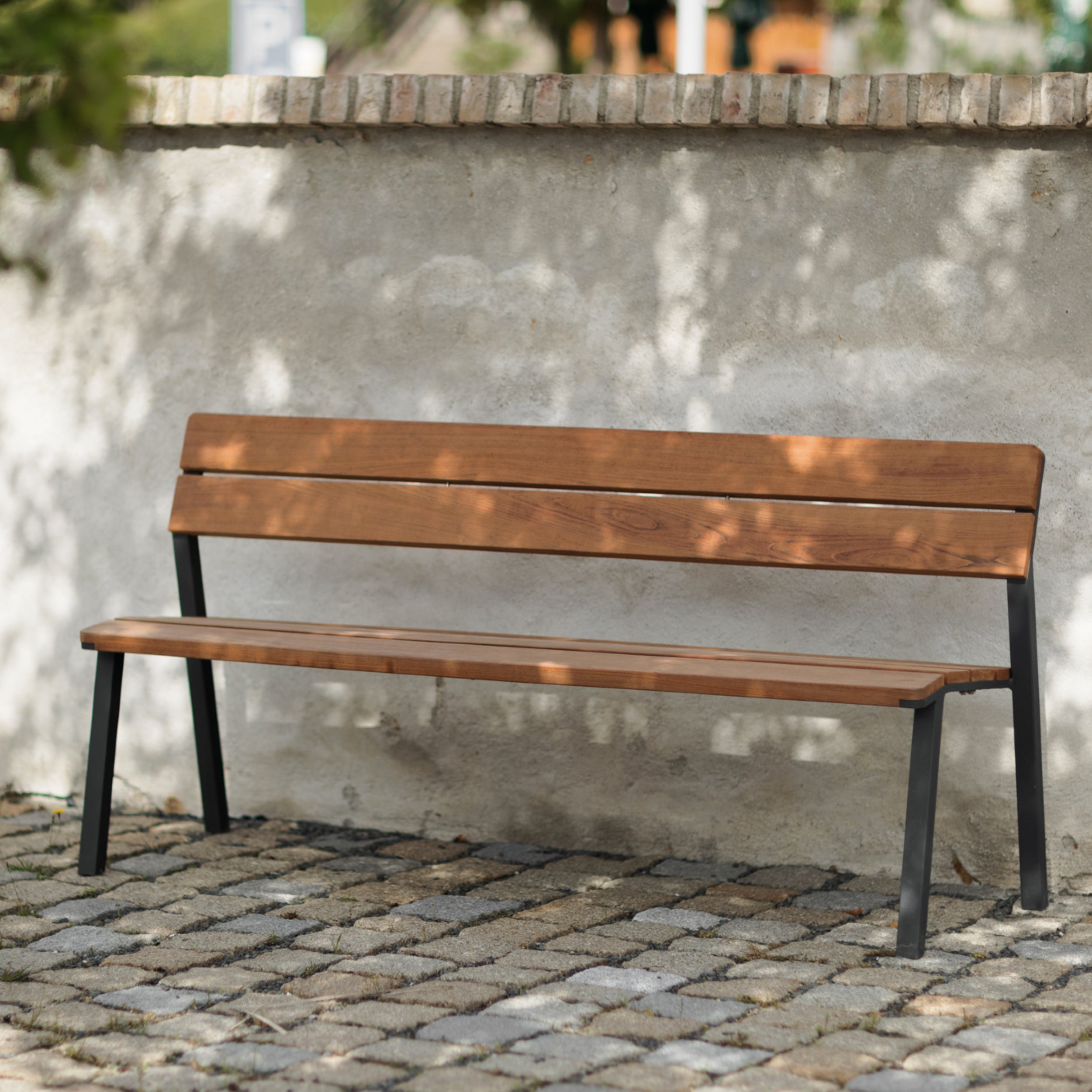The Cora Public Bench by Egoé, crafted from tropical Jatoba wood with a powder-coated steel frame, stands elegantly on a cobblestone surface against a beige brick wall. Sunlight filters through trees, creating dappled shadows on this refined piece of street furniture.
