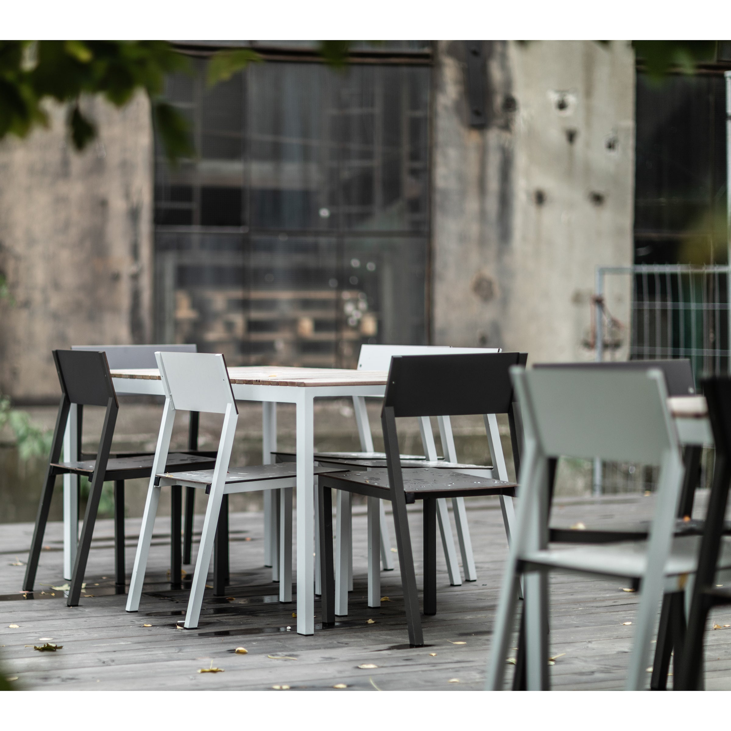 An outdoor seating area features sleek Cora Chairs by Egoe, a modern table, and six stackable chairs on a wooden deck. The industrial-style building in the background has large windows and visible wear, framed by lush greenery.