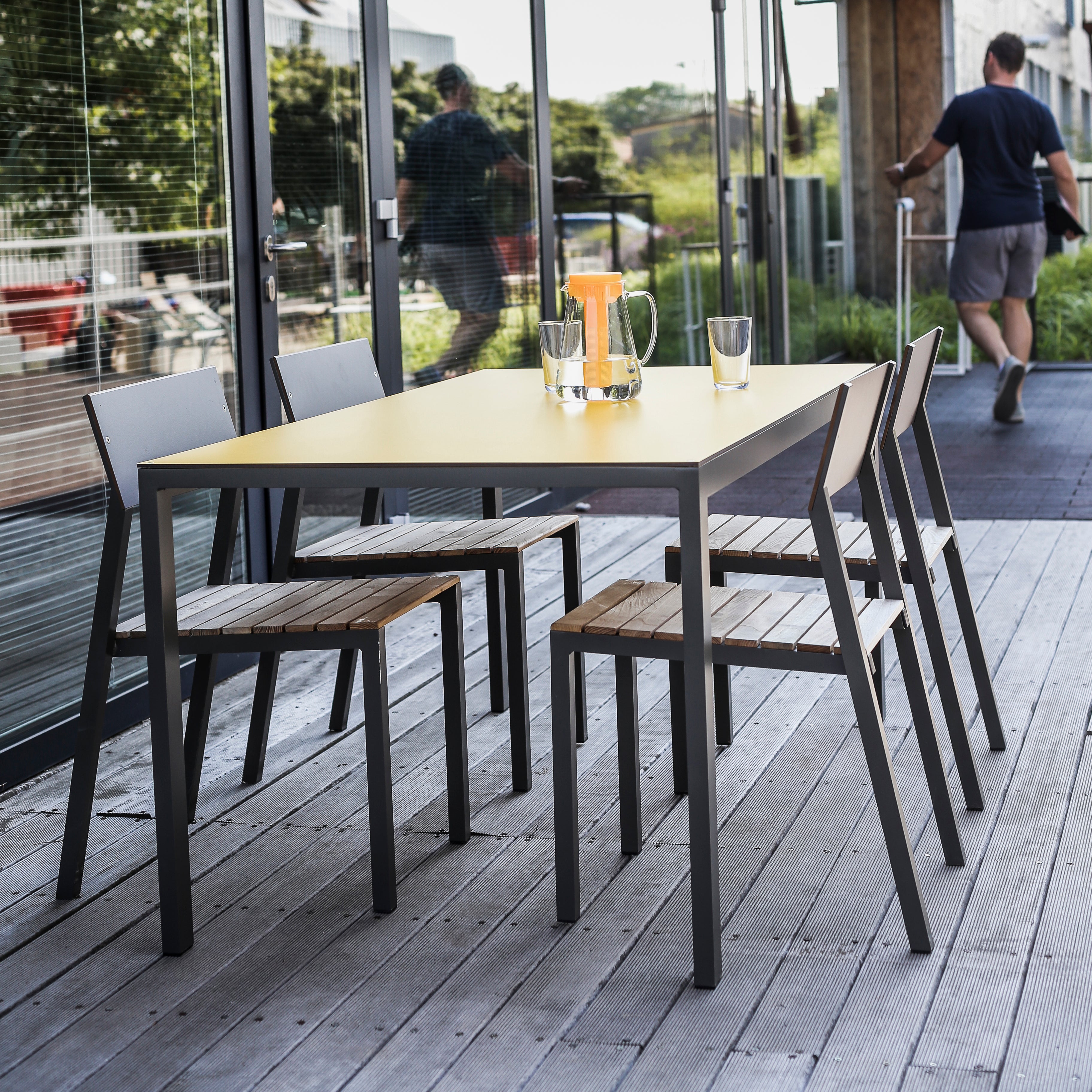 A modern outdoor dining area showcases a rectangular yellow table and sleek Egoe Cora chairs with wooden slats. A glass pitcher of orange juice with two glasses invites relaxation, while a passerby highlights the charm of stackable outdoor furniture beside a glass-walled building.