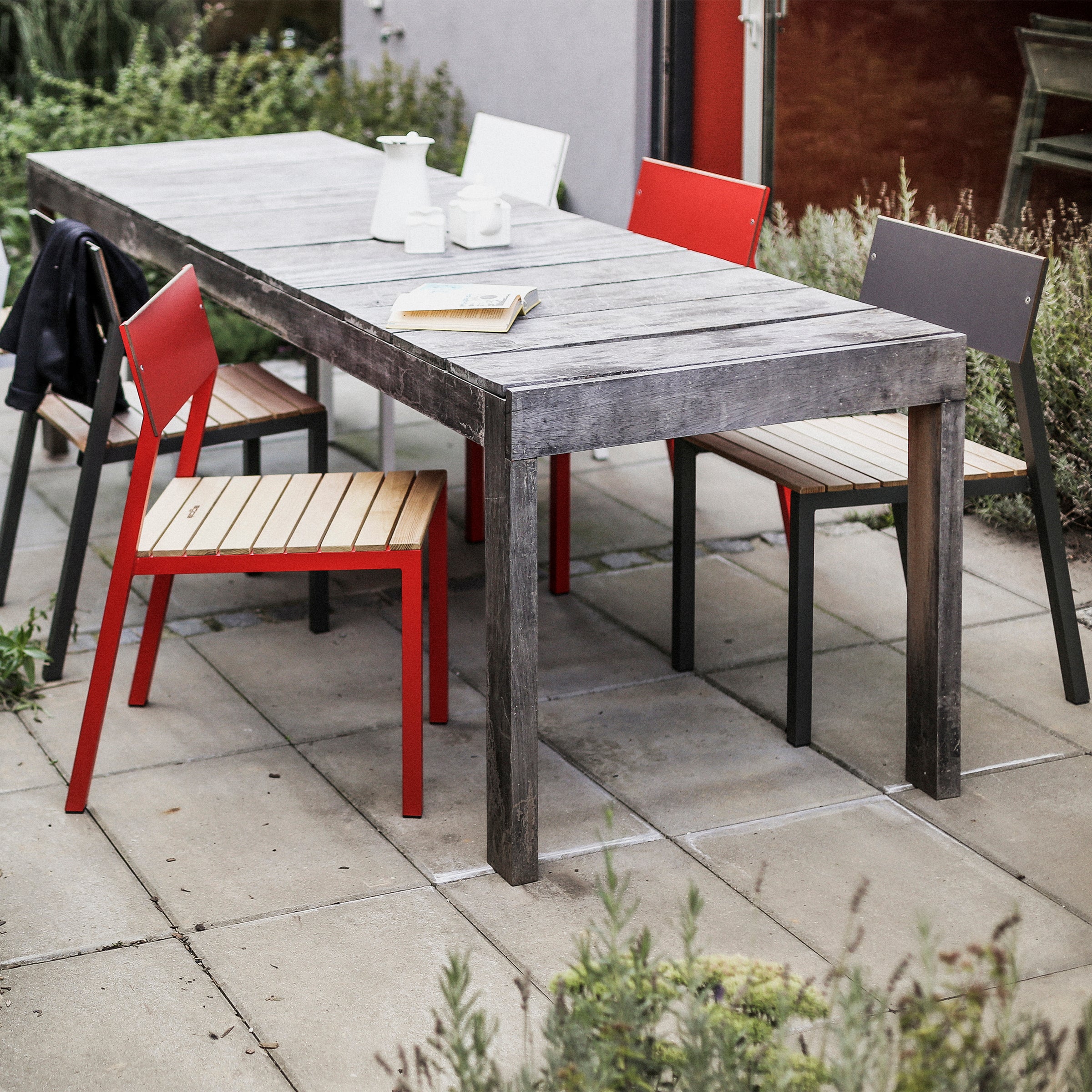 A wooden table with a gray weathered finish sits on a patio with gray tiles, surrounded by outdoor furniture, including red chairs and Egoe Cora Chairs. A white teapot, cups, a book, and a black jacket are on the table. Greenery adds to the inviting atmosphere.