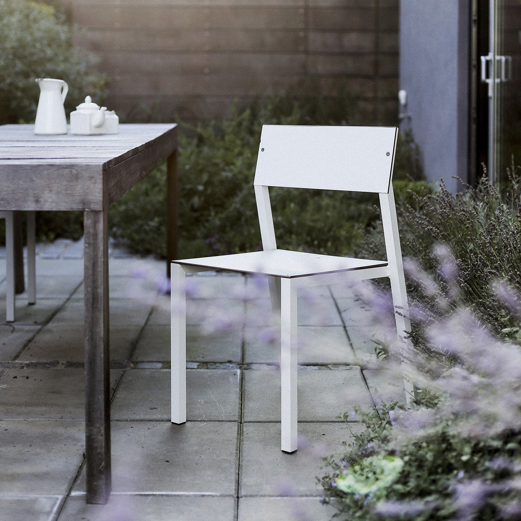 A minimalist Cora Chair by Egoe is set beside a wooden table on a stone patio. A small white teapot and creamer rest on the table, with blooming lavender in the foreground enhancing this elegant piece of outdoor furniture, as wooden paneling appears in the background.