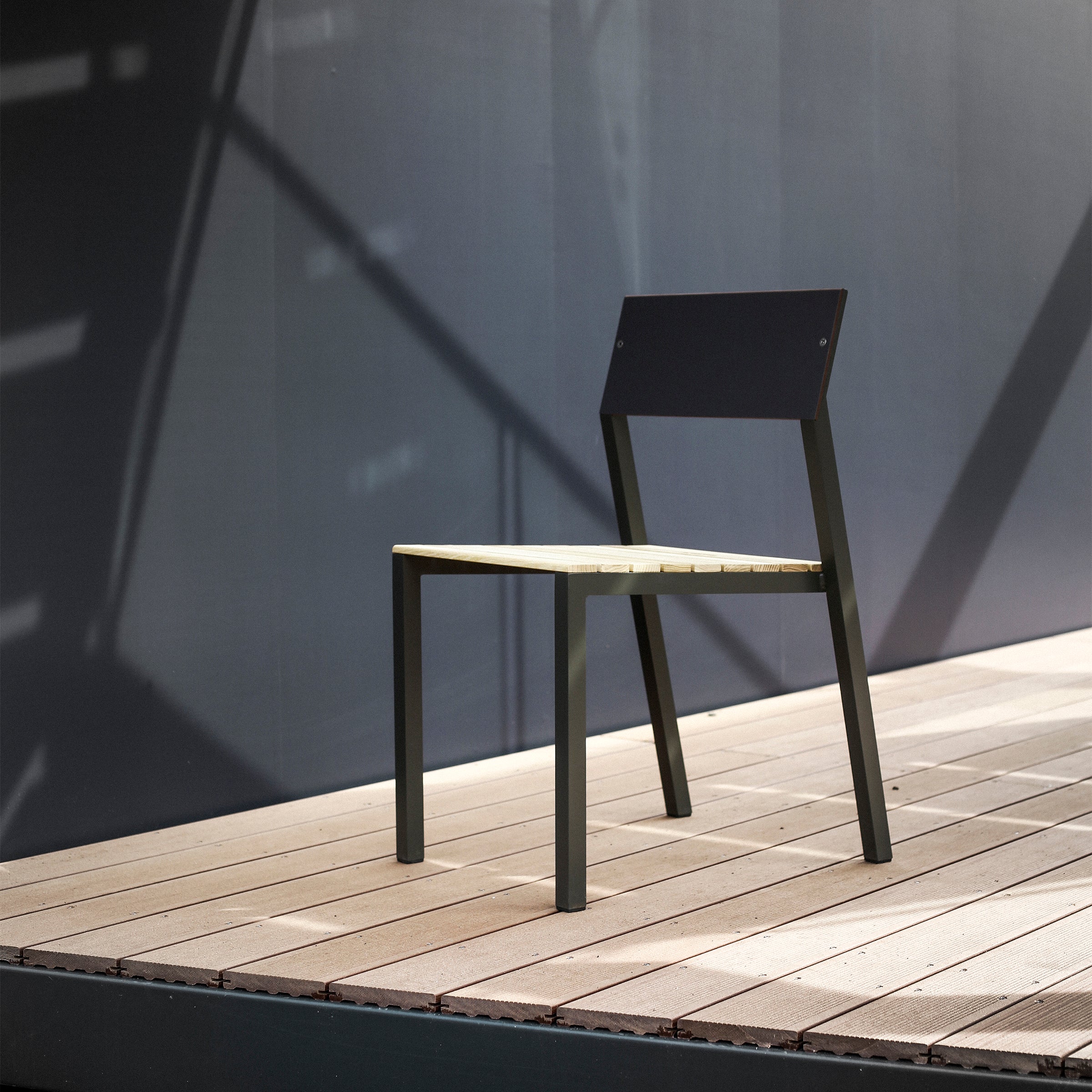 The minimalist Cora Chair by Egoe features a black backrest with light wood slats for the seat, set on a wooden deck. Positioned against a plain, dark wall, its design is highlighted by shadows from the railing.