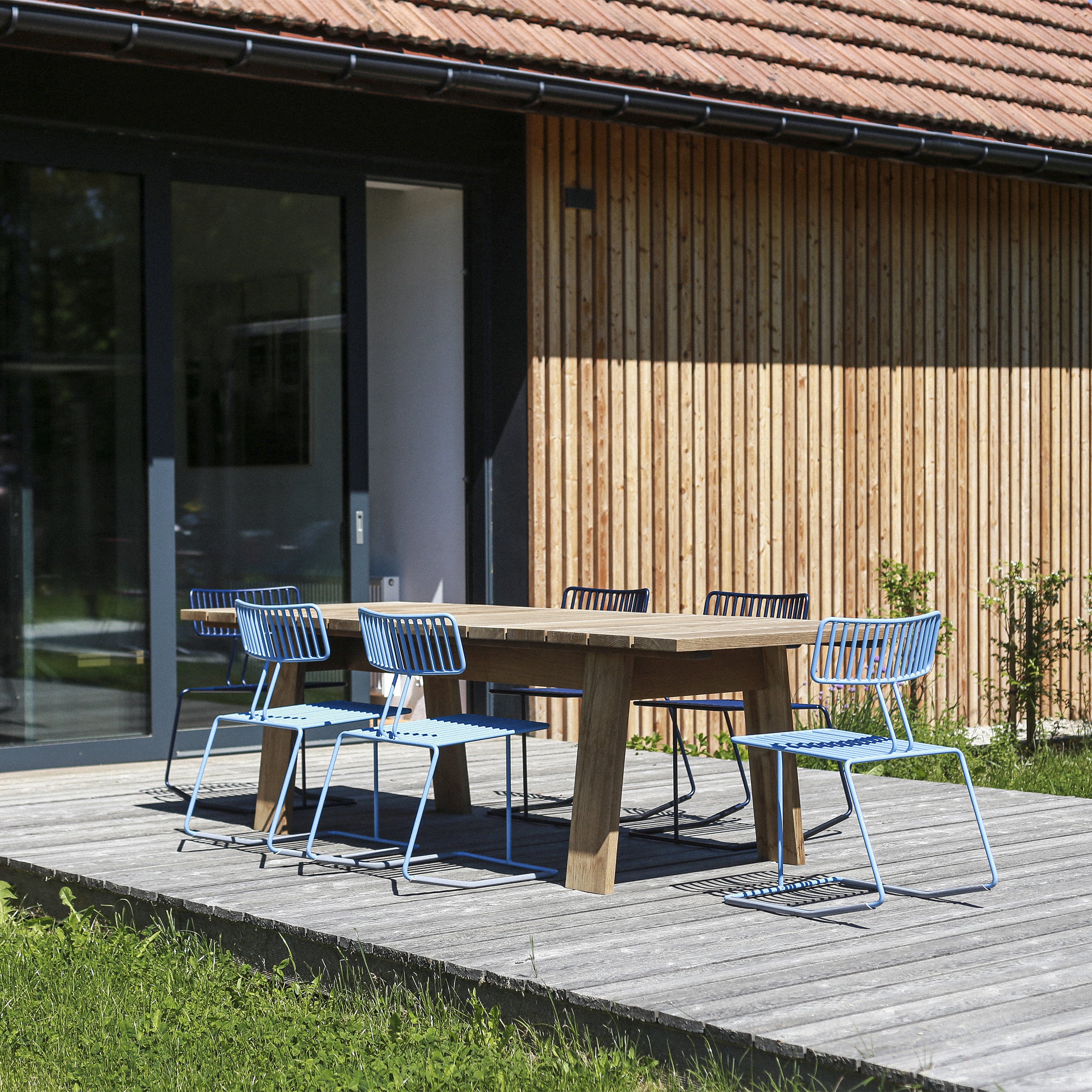 An outdoor patio features a wooden table and six elegant Hope Chairs by Egoé on a wooden deck. A modern house with vertical wood panels and large glass doors provides the backdrop, surrounded by a lush green lawn that enhances the serene atmosphere.