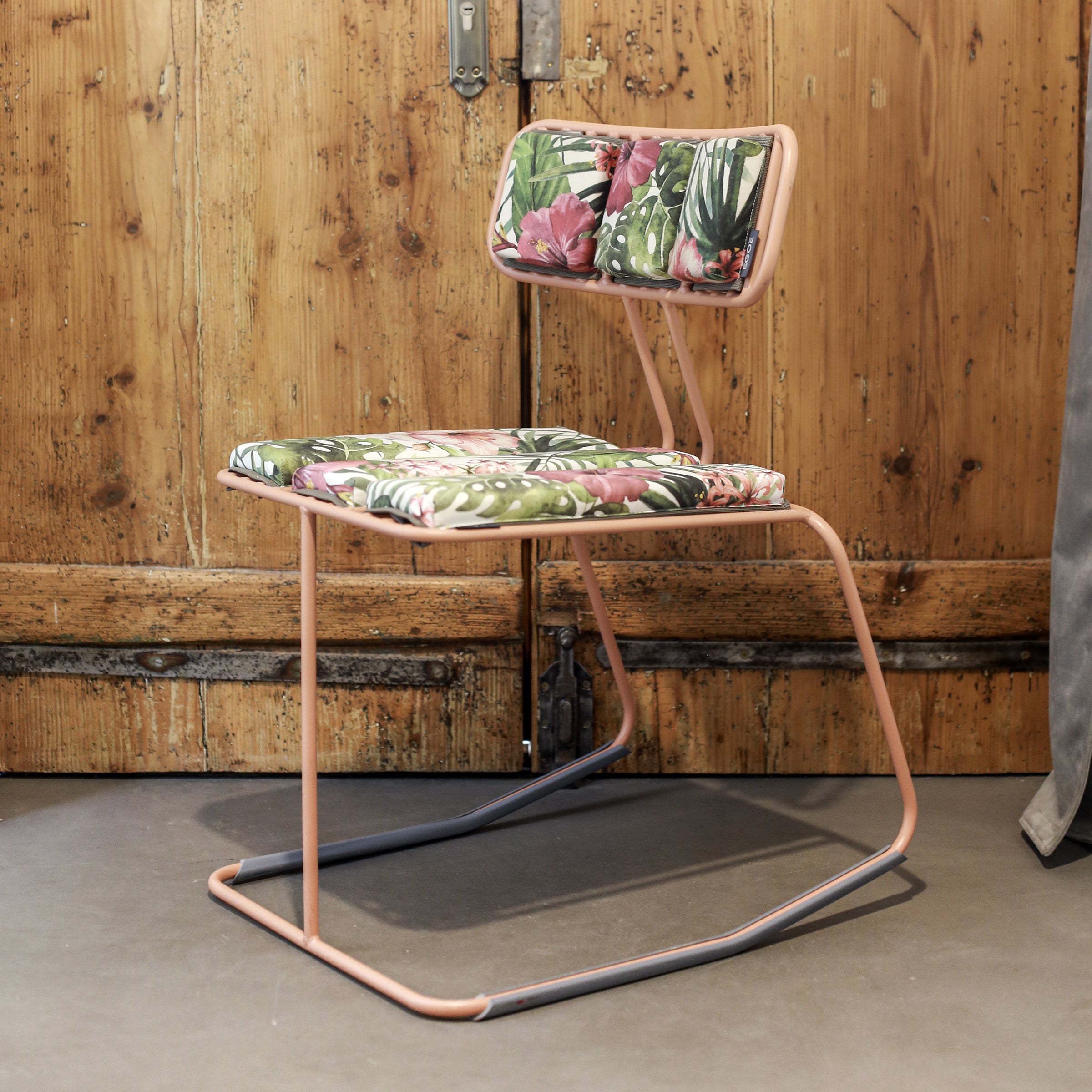 Egoés Hope Chair features a light pink metal frame and a cushion with green leaves and pink flowers, elegantly placed on a gray floor against a rustic wooden door.