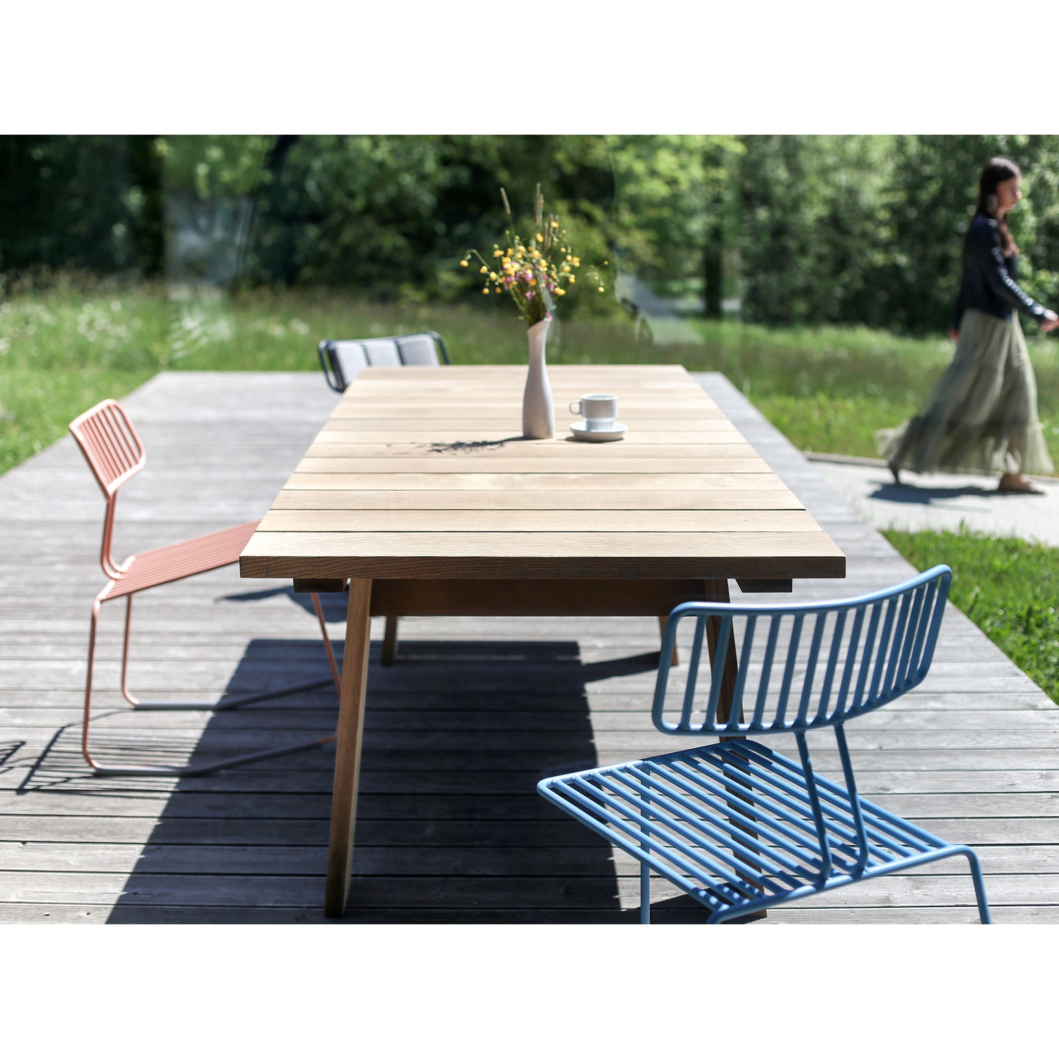 A cozy wooden outdoor table sits on a wooden deck with an Egoe Hope Chair - SALE and another metal chair in blue and pink. A vase with flowers and a cup adorn the table, while someone walks by amidst lush greenery in the background.