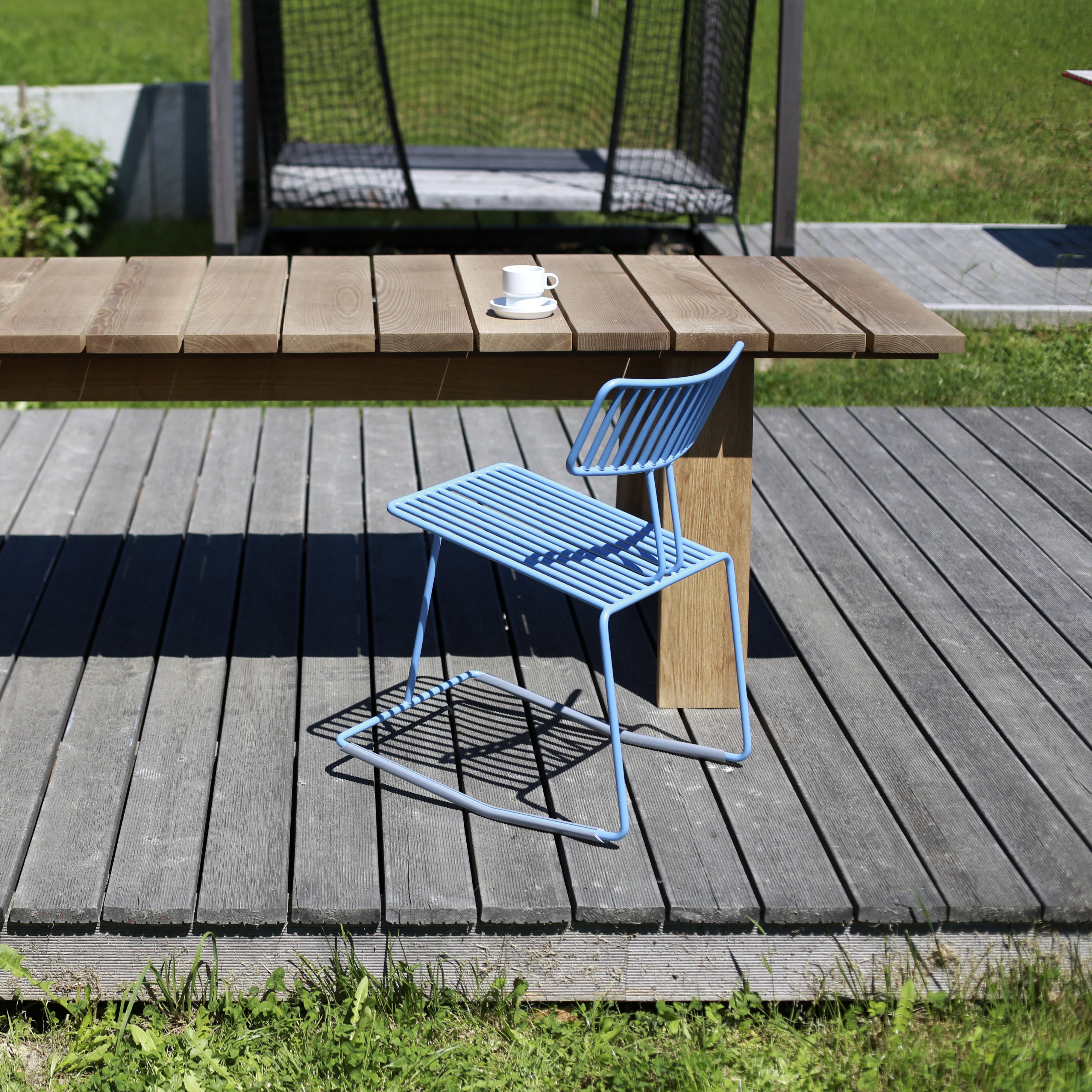A stylish Hope Chair by Egoé, with blue metal craftsmanship, adorns a wooden patio next to a matching table. A cup and saucer sit on the table, framed by vibrant green grass and the sleek lines of a modern pergola.