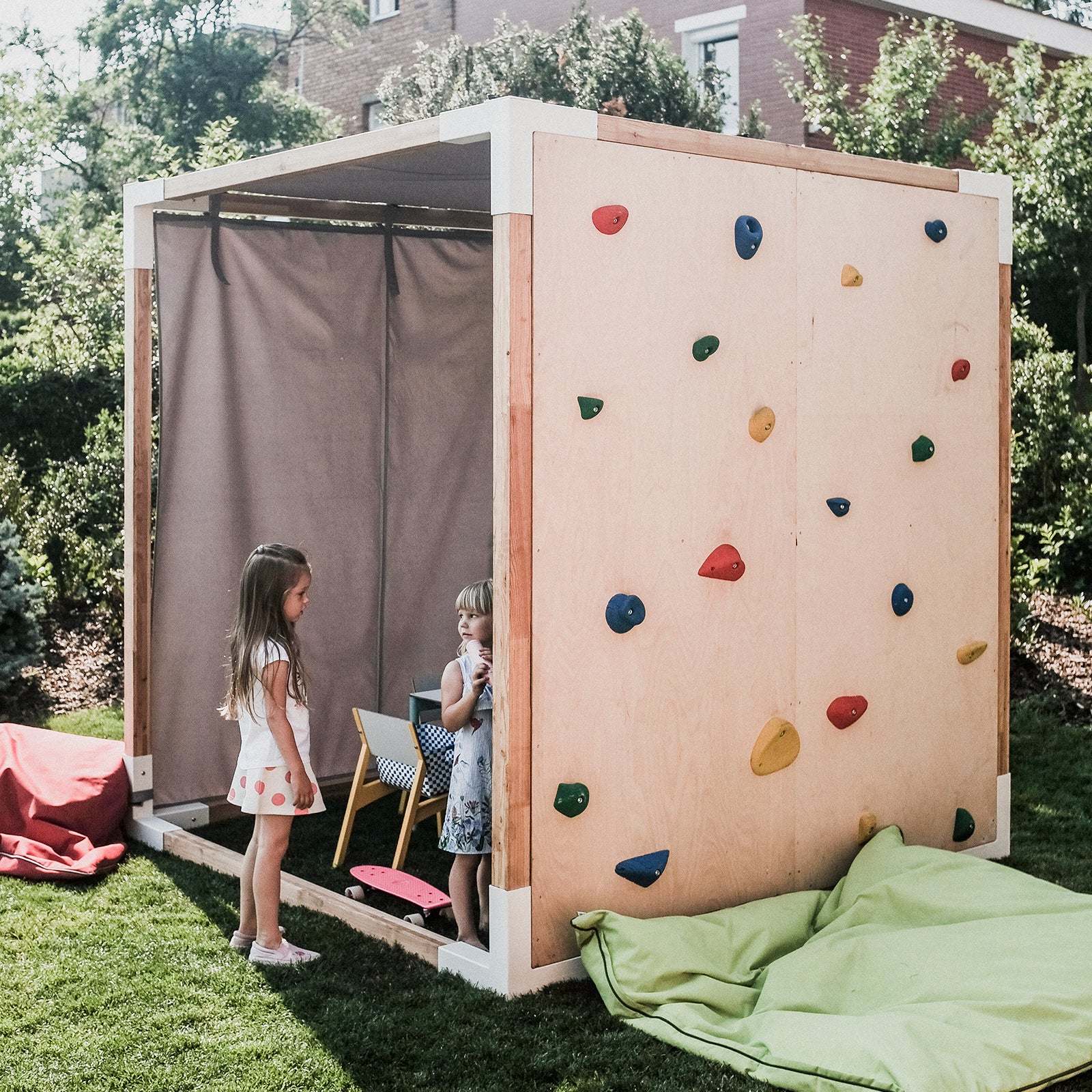 Two children stand by the Leva Home wooden play structure from Egoe, featuring colorful climbing holds. A partially enclosing curtain adds a sense of relaxation and tranquility. Green bean bags and red mats sit on the grass, framed by trees and a brick building.