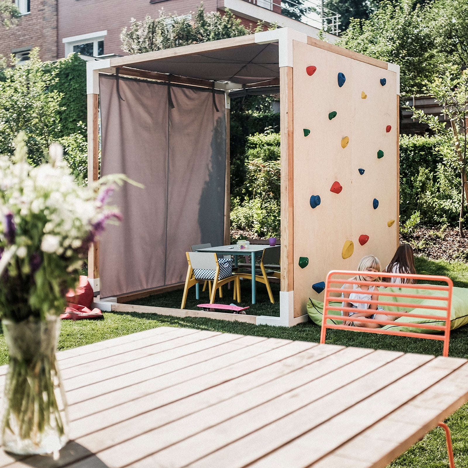 In the serene backyard of Leva Home by Egoe, elegant outdoor furniture graces the scene: a wooden table with flowers, a climbing wall by a shelter with seating, all surrounded by greenery where two children play near distant brick buildings.