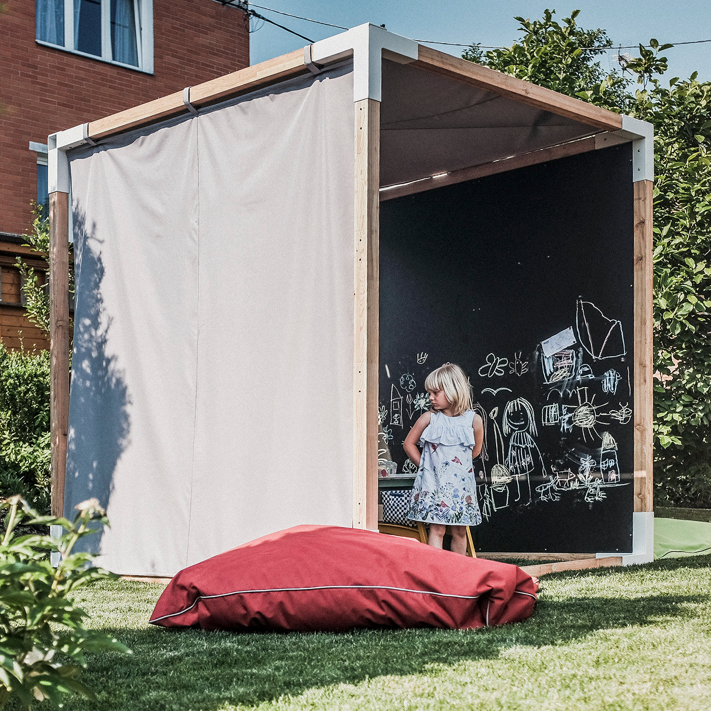 In a backyard by a brick building, a child in a floral dress enjoys the Egoe Leva Home playhouse with its black chalkboard wall full of drawings. Nearby, outdoor furniture and a large red cushion rest on the grass, offering relaxation under the shade of a cream curtain.
