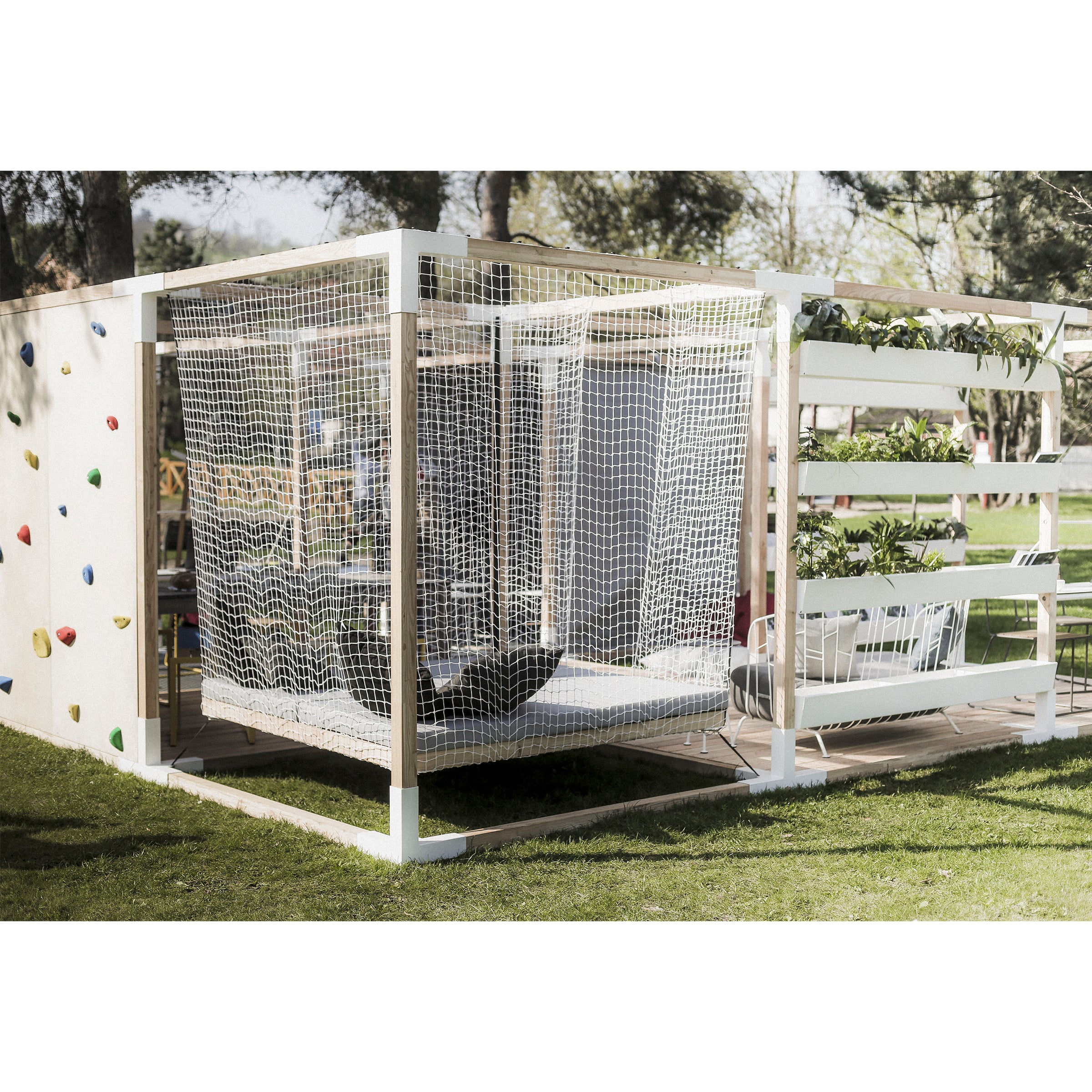 Leva Home by Egoe is a modern outdoor play area with a climbing wall, suspended nets, vertical planter boxes, and elegant outdoor furniture. Set in a grassy park with trees, sunlight filtering through creates a tranquil and relaxing atmosphere.