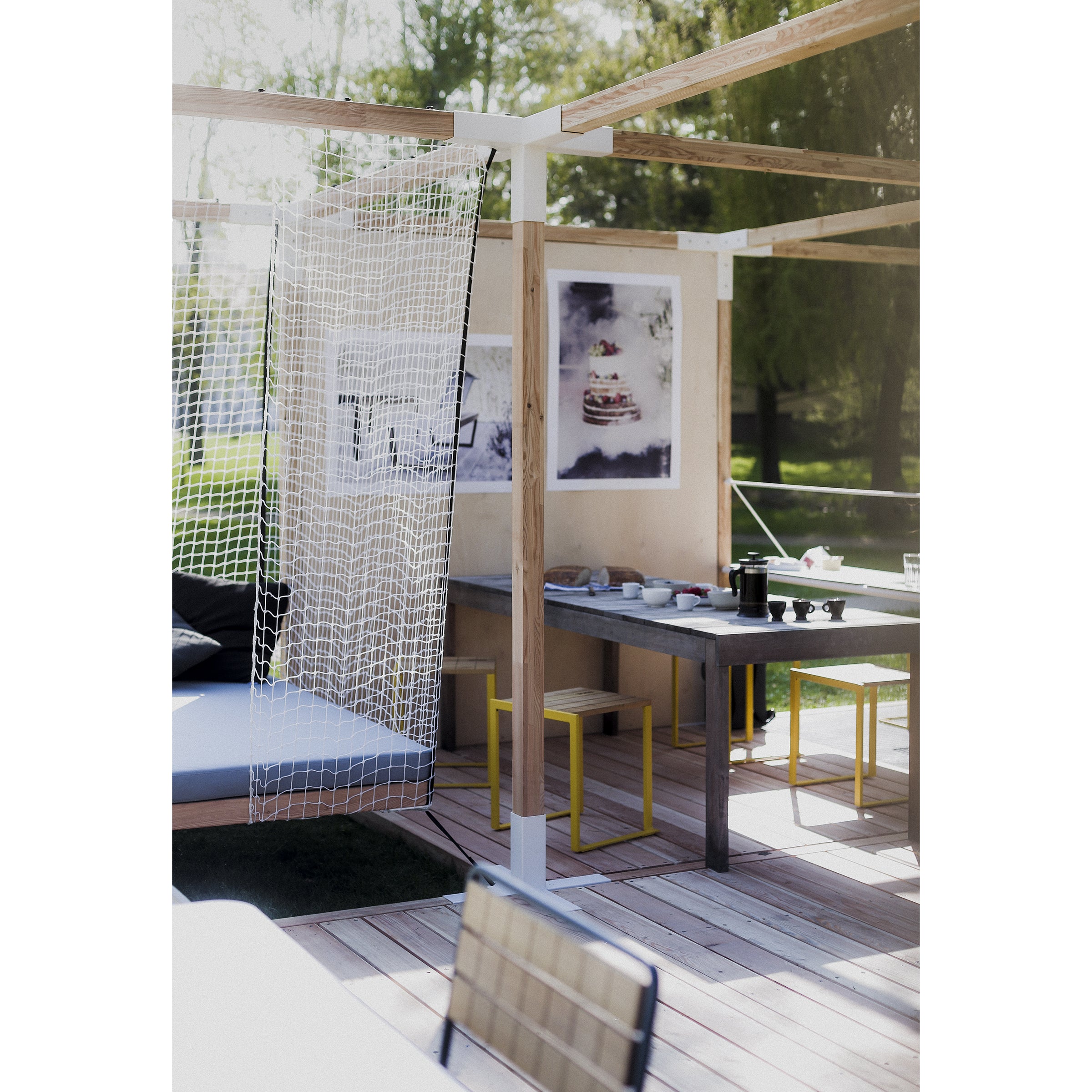 A modern outdoor patio with a pergola features Egoés Leva Home collection, including a wooden dining table set and framed artwork. The netted hammock adds elegance, while the lush greenery creates a cozy and inviting atmosphere.