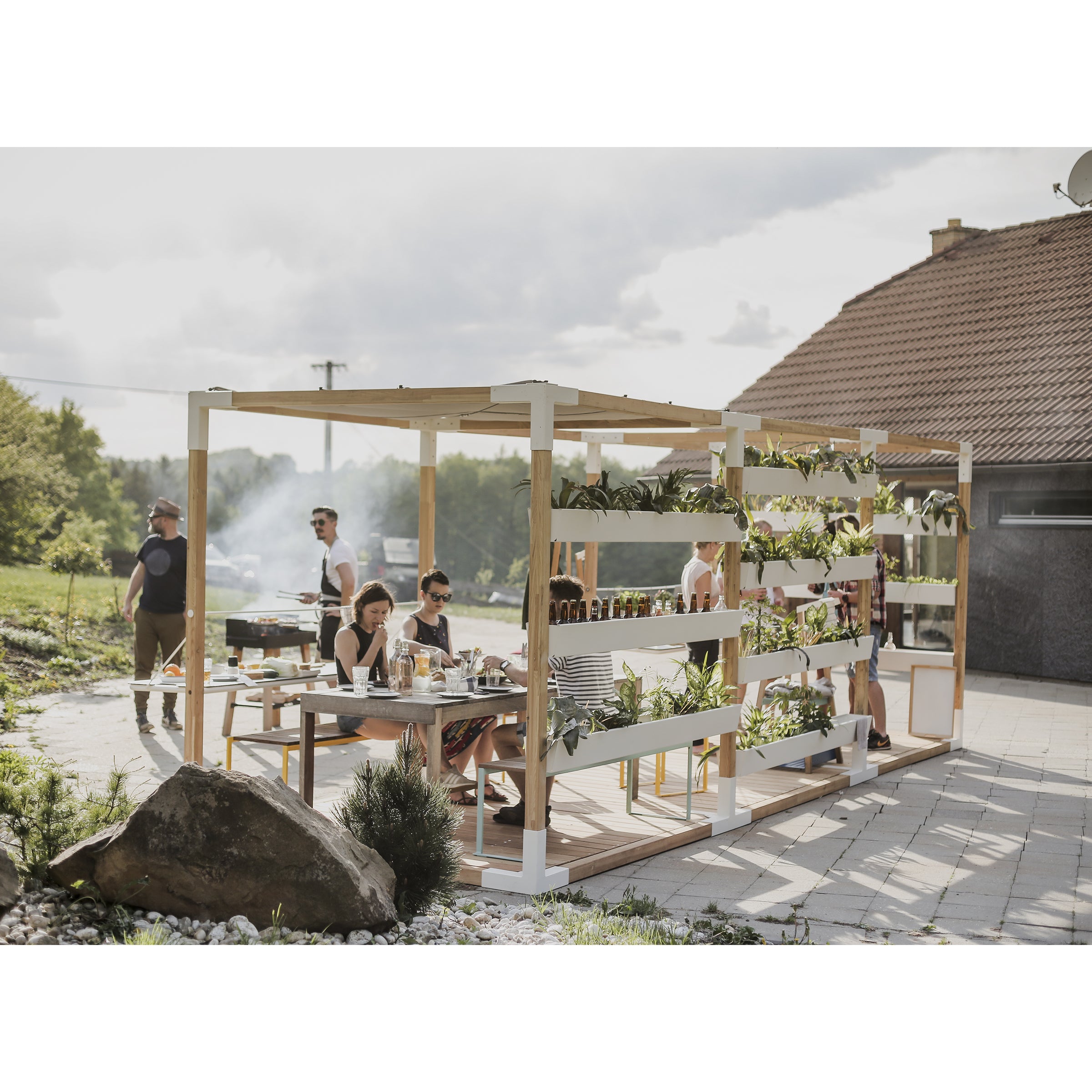 People are enjoying a meal under the Leva Home wooden pergola by Egoe on a patio, surrounded by outdoor furniture. The pergola features potted plants on shelves, and someone nearby is grilling. The scene is serene with a house and lush greenery in the background.