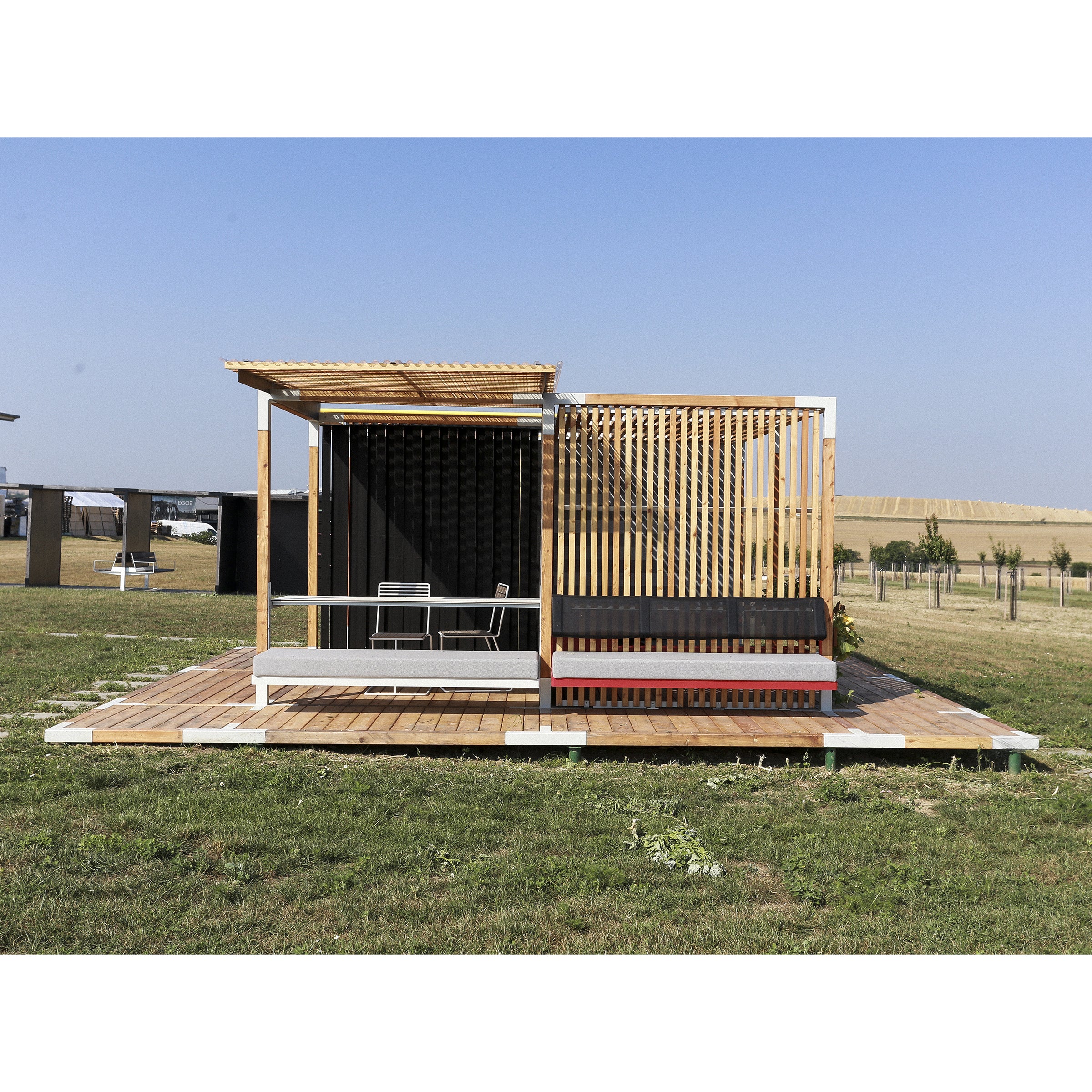 A modern, minimalist tiny house with a wooden facade and large windows features an Egoe Leva Office system-inspired slatted sunshade. Situated on a grassy lawn under a clear blue sky, it offers a small deck with outdoor seating, ideal for converting into an outdoor office space.