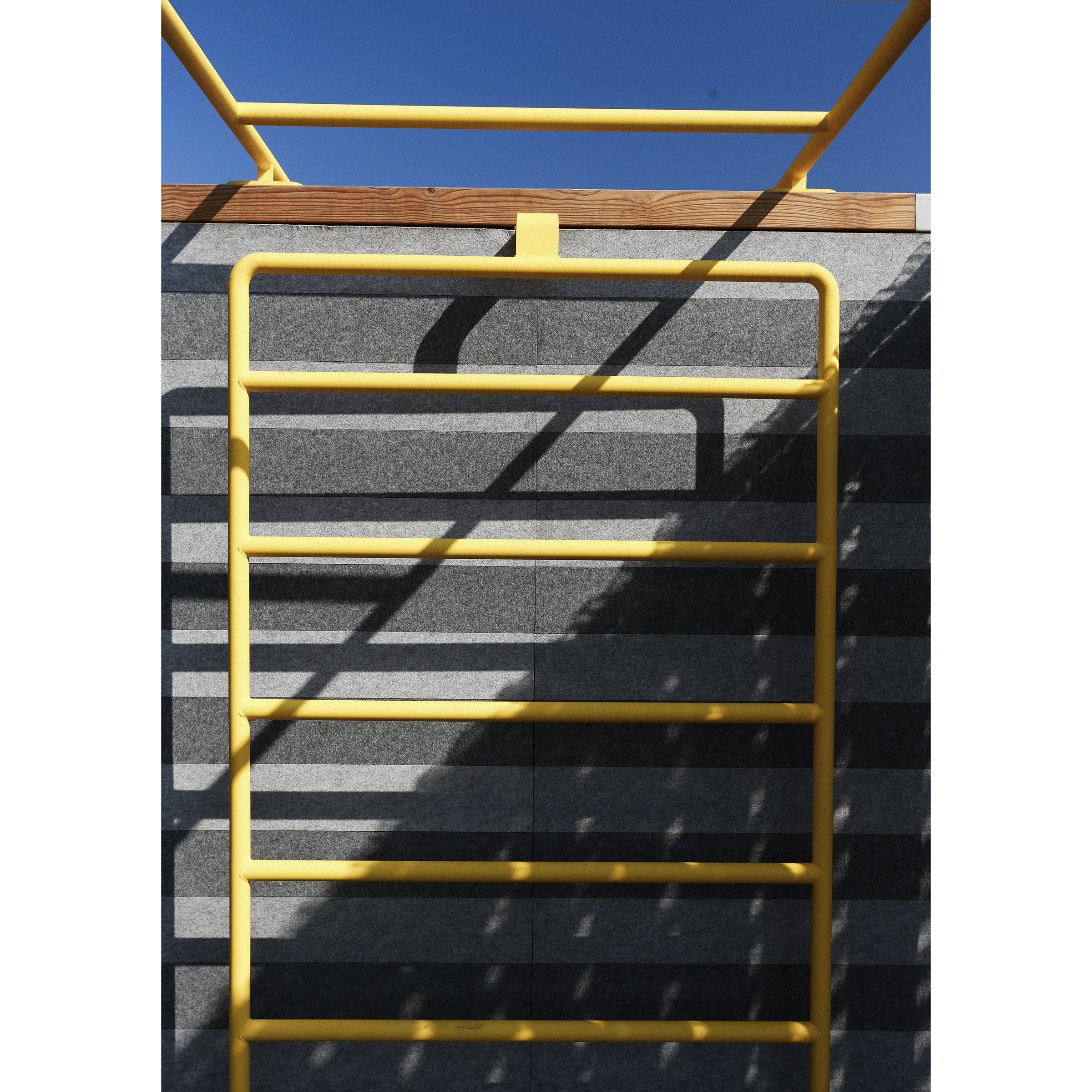The Egoé Leva Office and Classroom ladder leans against the gray brick wall, casting intricate shadows under a wide blue sky, suggesting an outdoor office setting infused with creativity and open-air inspiration.