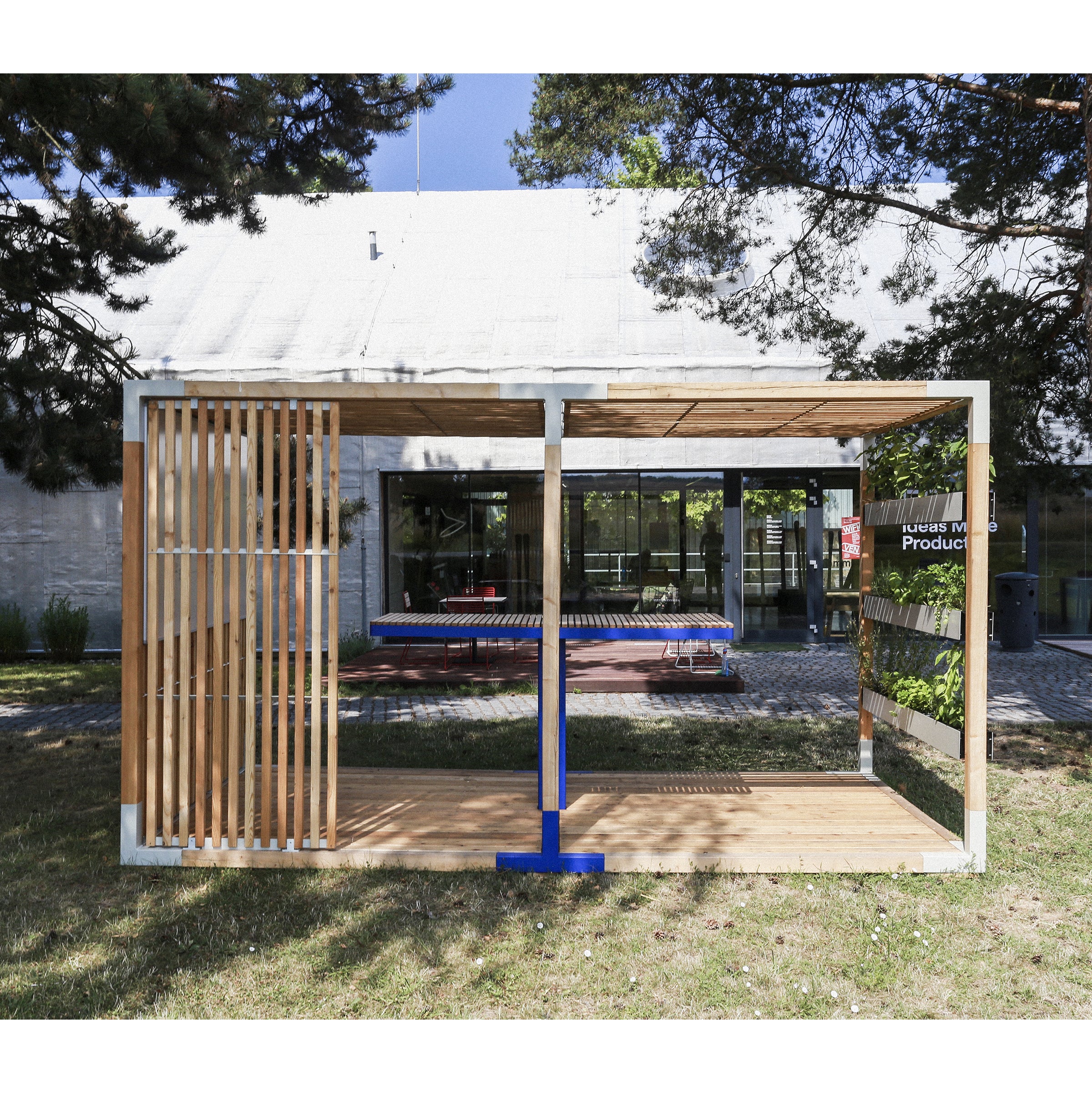 The Egoé Leva Office and Classroom showcases a modern design with wooden slats and a blue beam. Positioned on a grassy lawn before glass doors, it features the Leva system for minimalism while trees frame the area, casting shadows on the grass.