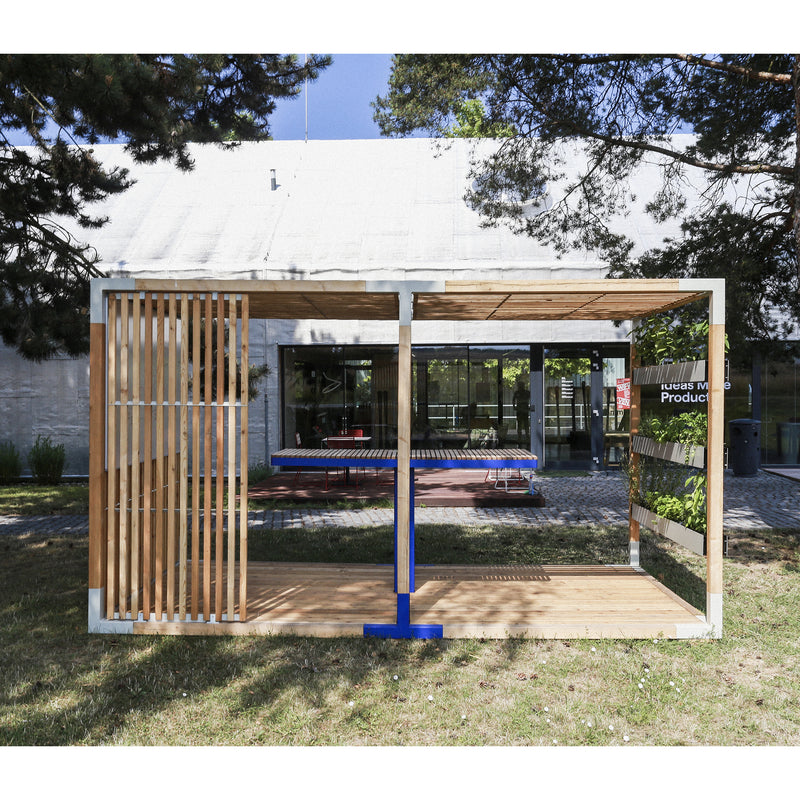 The Egoé Leva Office and Classroom showcases a modern design with wooden slats and a blue beam. Positioned on a grassy lawn before glass doors, it features the Leva system for minimalism while trees frame the area, casting shadows on the grass.
