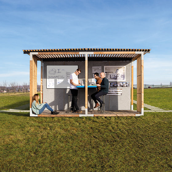 A modern outdoor workspace using the Leva Office and Classroom by Egoé accommodates four people; two at a standing desk, two on stools. The wooden-framed structure with whiteboards stands on a grassy field beneath a bright blue sky.