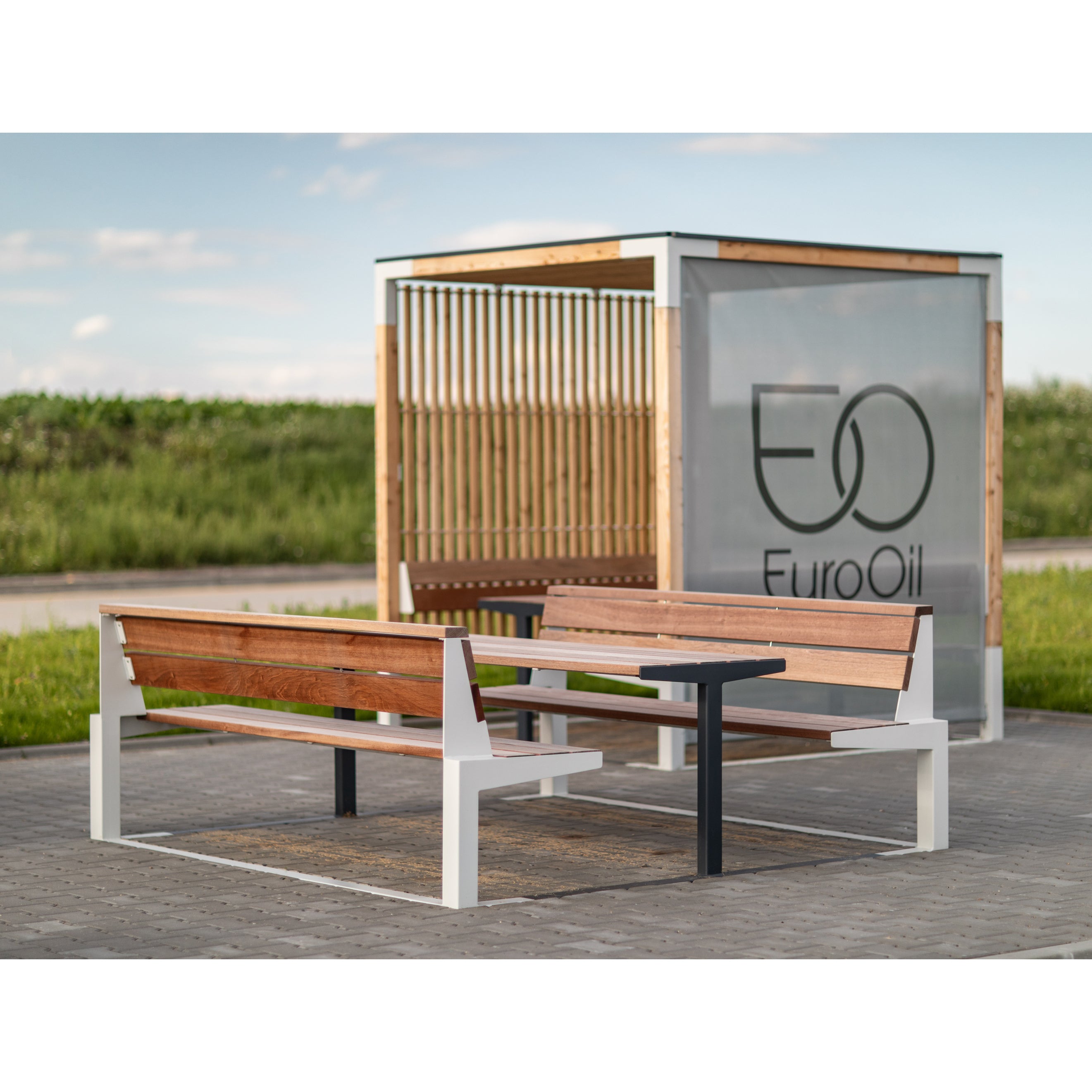 At the EuroOil station, the outdoor seating area serves as a relaxation zone with Leva Public wooden benches and table from Egoe on a paved surface. A wooden slatted structure with the EuroOil logo stands behind, surrounded by lush greenery under a partly cloudy sky.