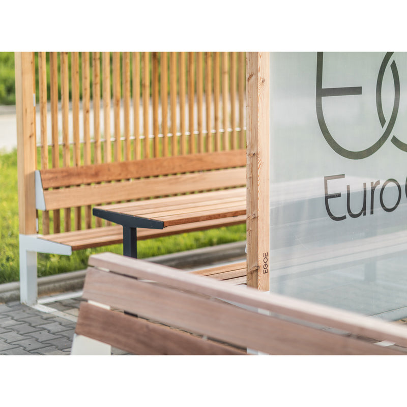 The Leva Public bench by Egoe features a wooden design with a glass panel displaying partial lettering, wrapped in a slatted structure for an outdoor office feel. Grey pavement and lush greenery in the background make it an inviting relaxation spot.