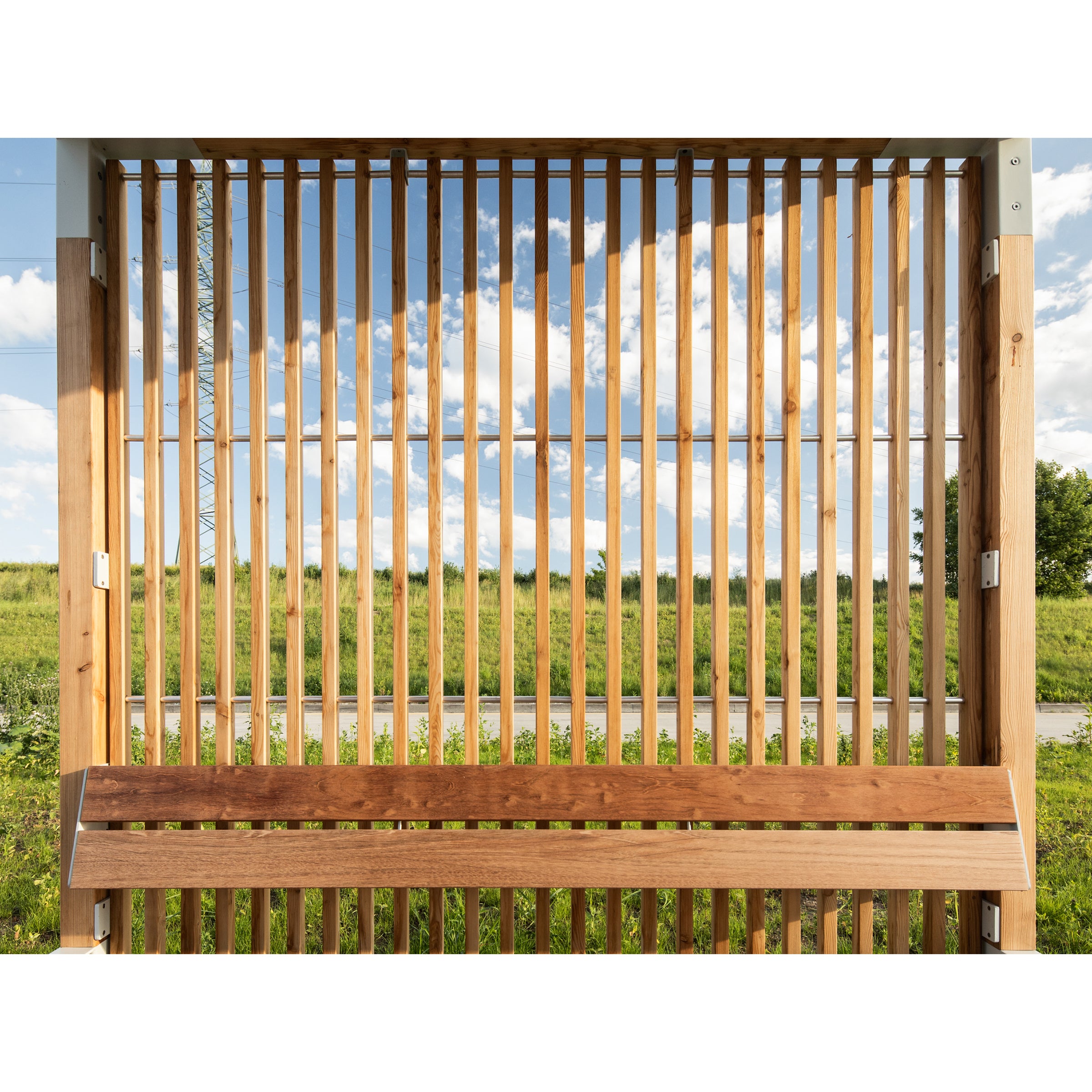 Vertical wooden slats of the Egoe Leva Public, with a transparent panel, frame green grass and a road under a blue sky with white clouds. This design is perfect for an outdoor office or relaxation space.