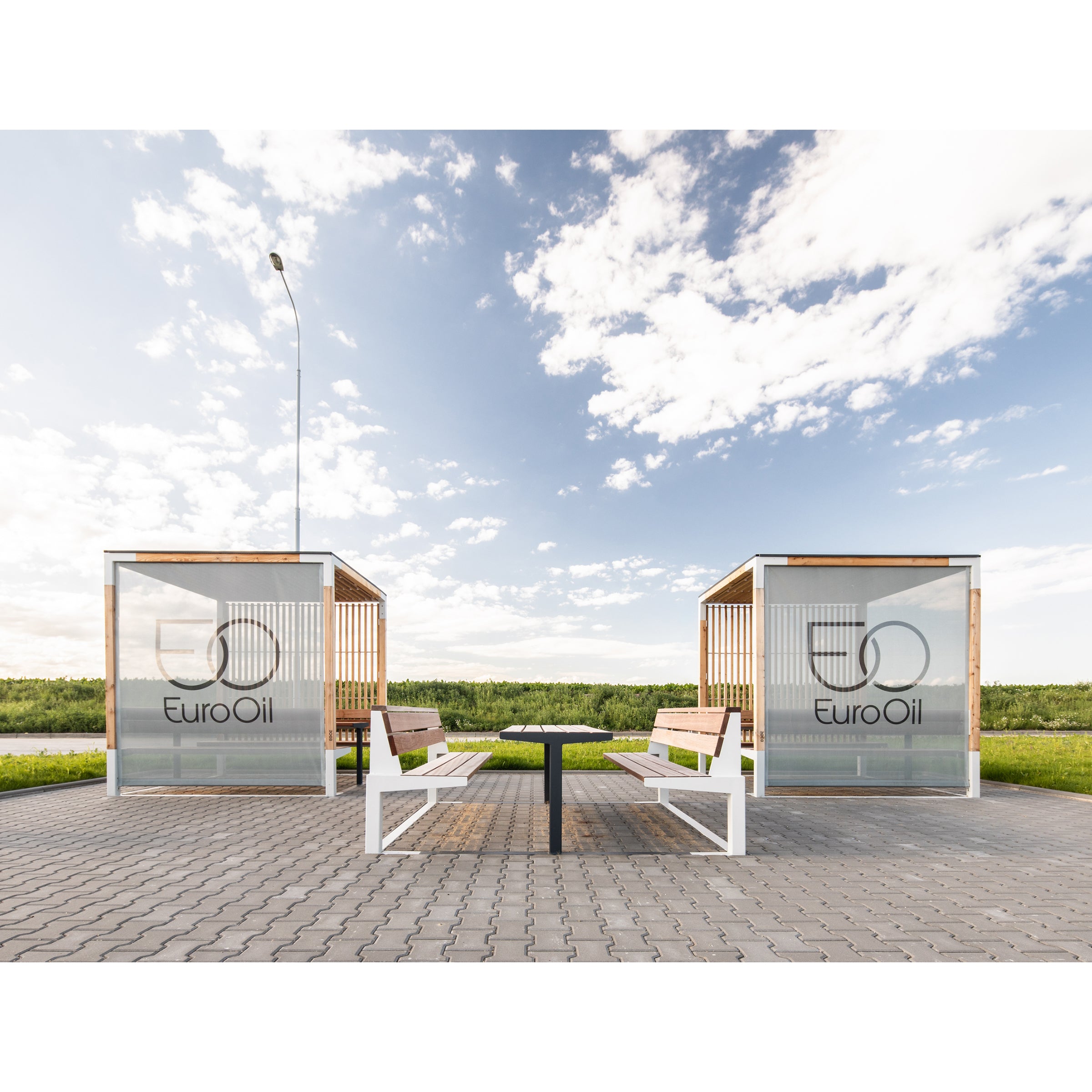 Two outdoor seating areas featuring Leva Public benches by Egoe offer a peaceful relaxation space against a vast green field and partly cloudy sky. Ideal for an outdoor office setup, these areas are sheltered by contemporary wooden and glass structures.