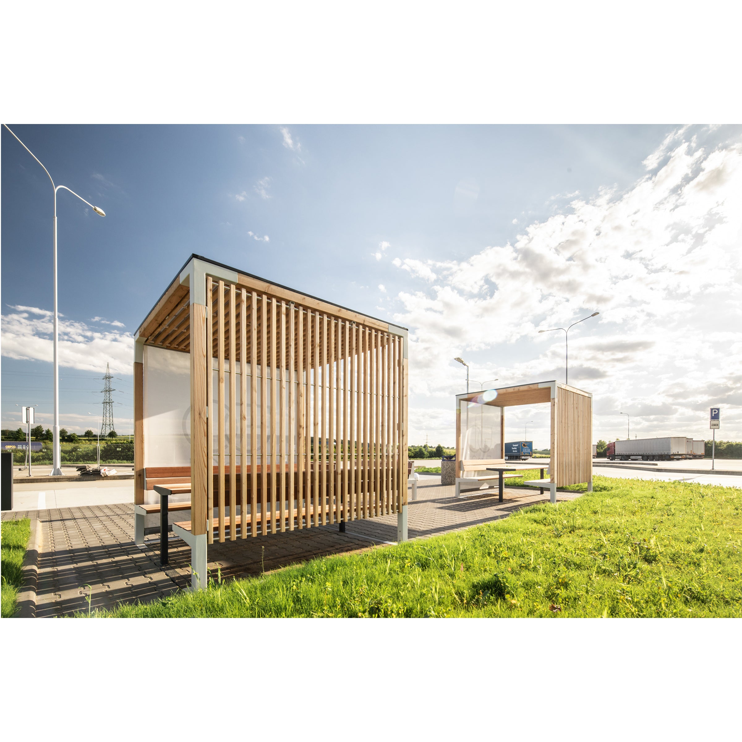 Two Egoe Leva Public outdoor bus shelters, featuring wooden slats and benches, offer an ideal relaxation spot on sunny days. They are placed on a paved area amidst green grass under a clear blue sky with scattered clouds.