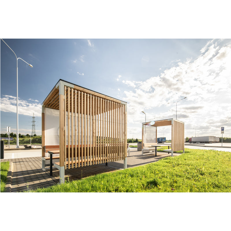 Two Egoe Leva Public outdoor bus shelters, featuring wooden slats and benches, offer an ideal relaxation spot on sunny days. They are placed on a paved area amidst green grass under a clear blue sky with scattered clouds.