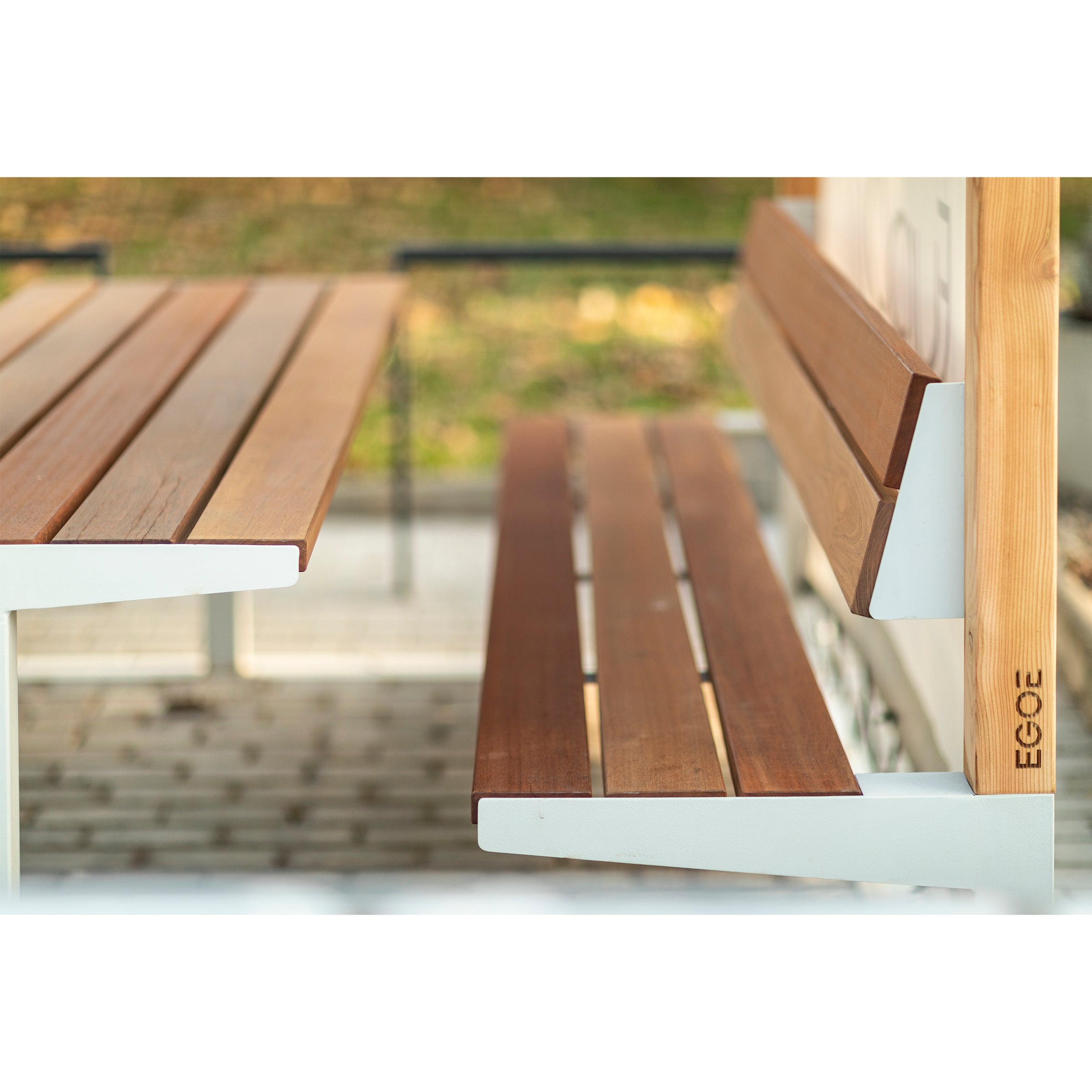 The Leva Public bench by Egoe features light brown wooden slats and a white metal frame, situated on a gray brick pathway with greenery in the background, enhancing this relaxation space.