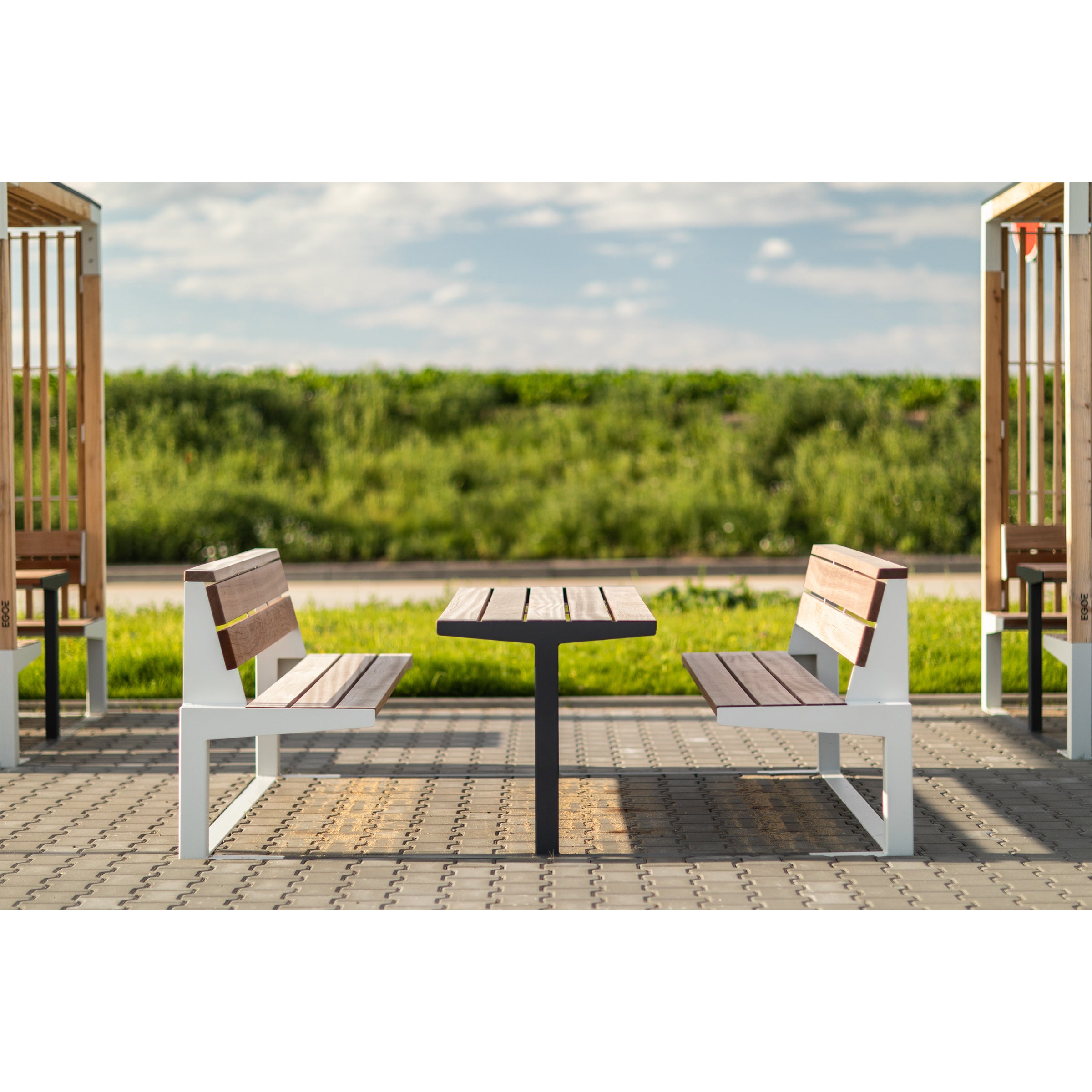 Two Leva Public wooden benches by Egoé face each other with a table in between on a paved area, against greenery and a cloudy blue sky, forming a serene outdoor office scene.