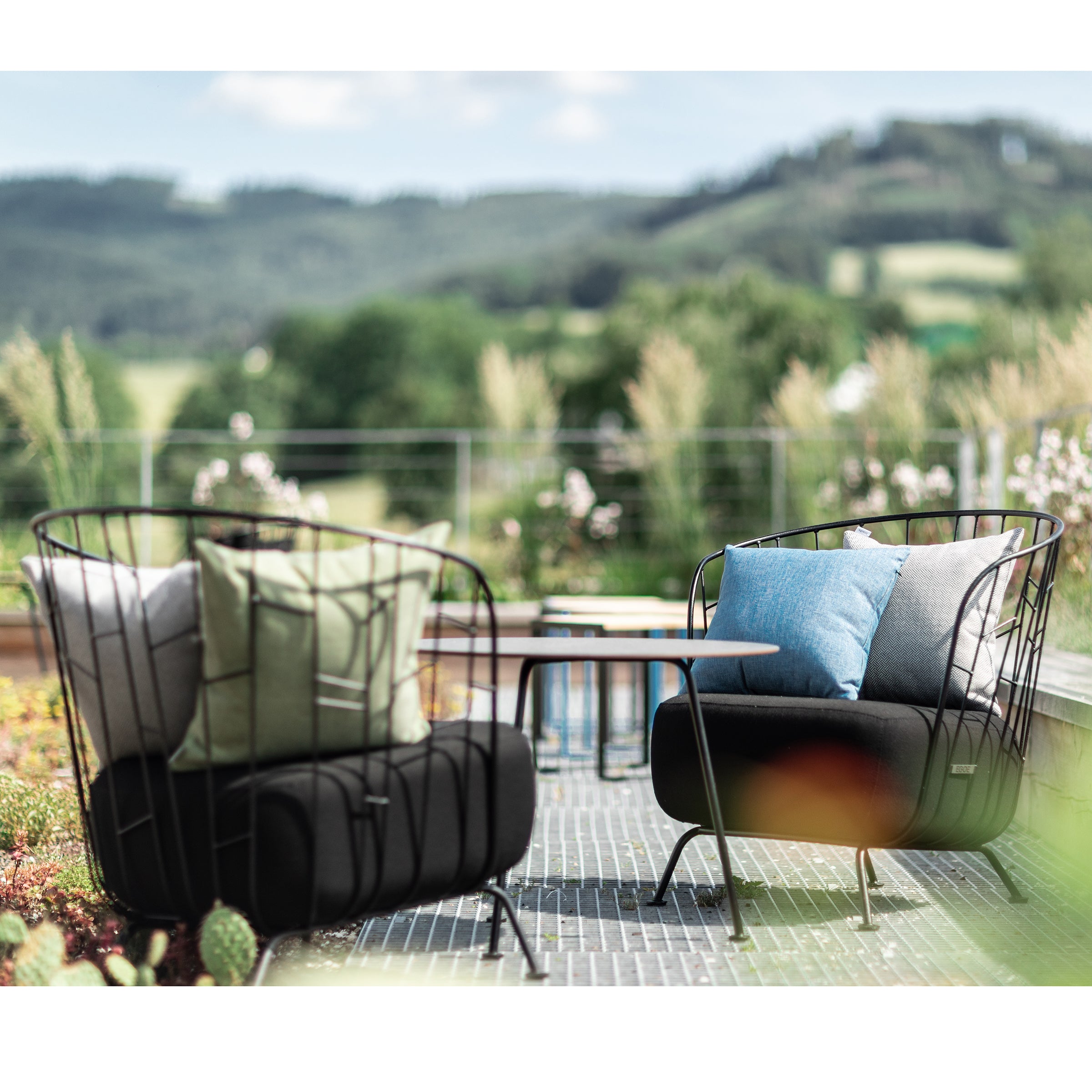 Two Maj Lounge Chairs by Egoe, made of zinc-coated steel with cushions, face each other on a patio table amidst scenic rolling green hills and trees under a clear blue sky.
