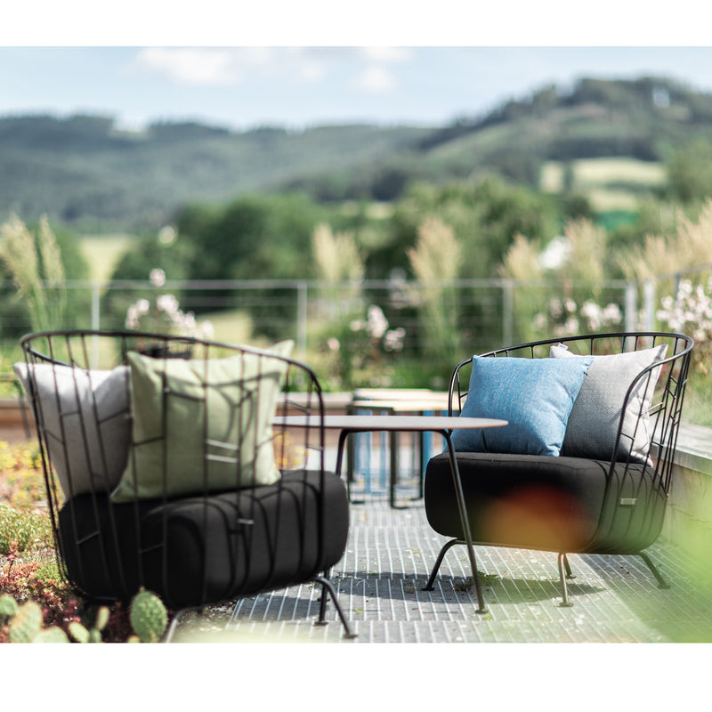 Two Maj Lounge Chairs by Egoe, made of zinc-coated steel with cushions, face each other on a patio table amidst scenic rolling green hills and trees under a clear blue sky.