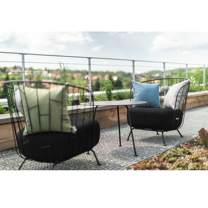 A Maj Lounge Chair in green pairs with a blue one from Egoe on a rooftop patio. Theyre next to a small high-pressure laminate table, all within zinc-coated steel glass railings that reveal distant greenery and partly cloudy skies.