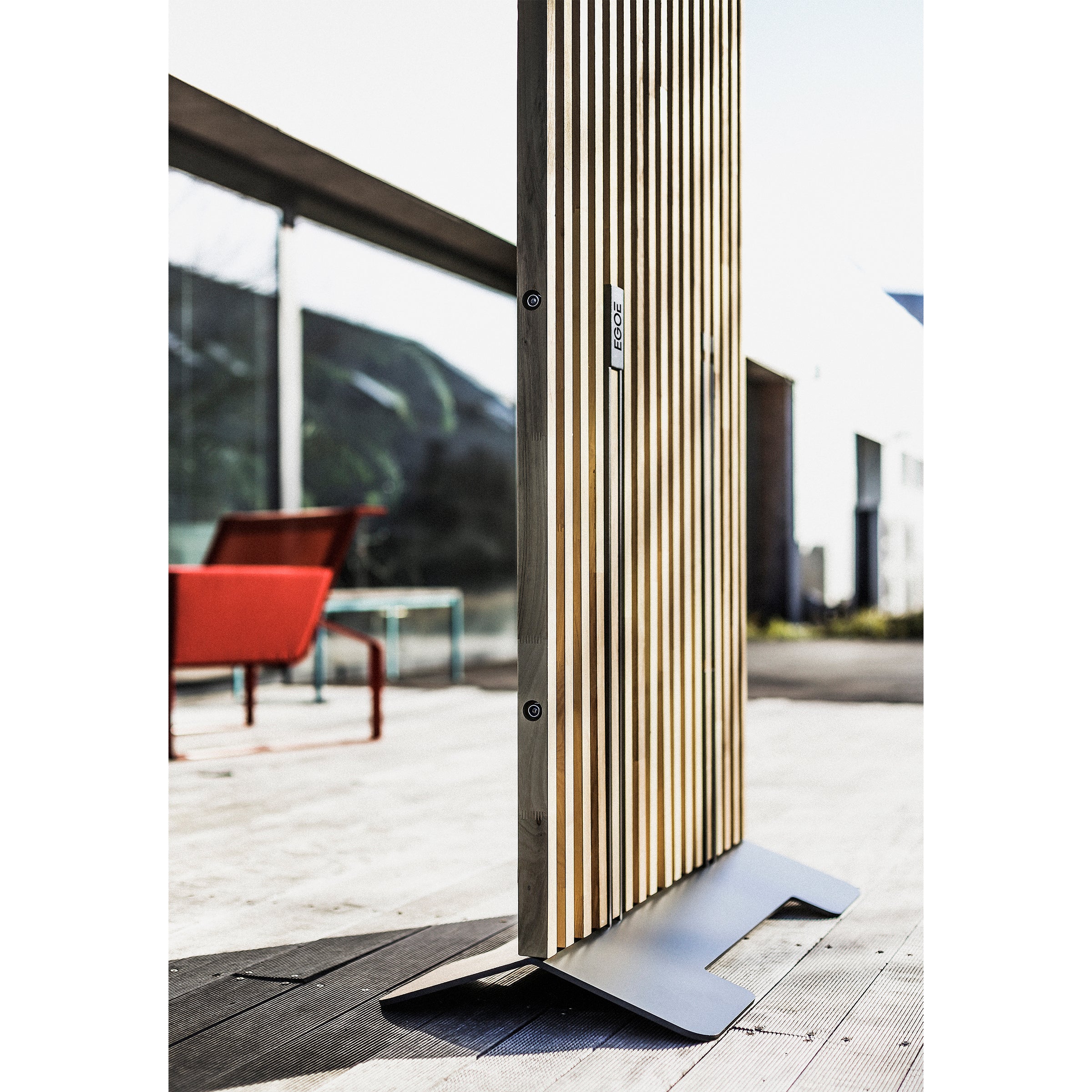 The Mario Screen by Egoe, a wooden slatted outdoor panel with a galvanized steel base, adorns the deck. Nearby, a red chair and glass table complement the modern architecture and greenery, providing style and privacy under a clear sky that enhances the tranquil setting.