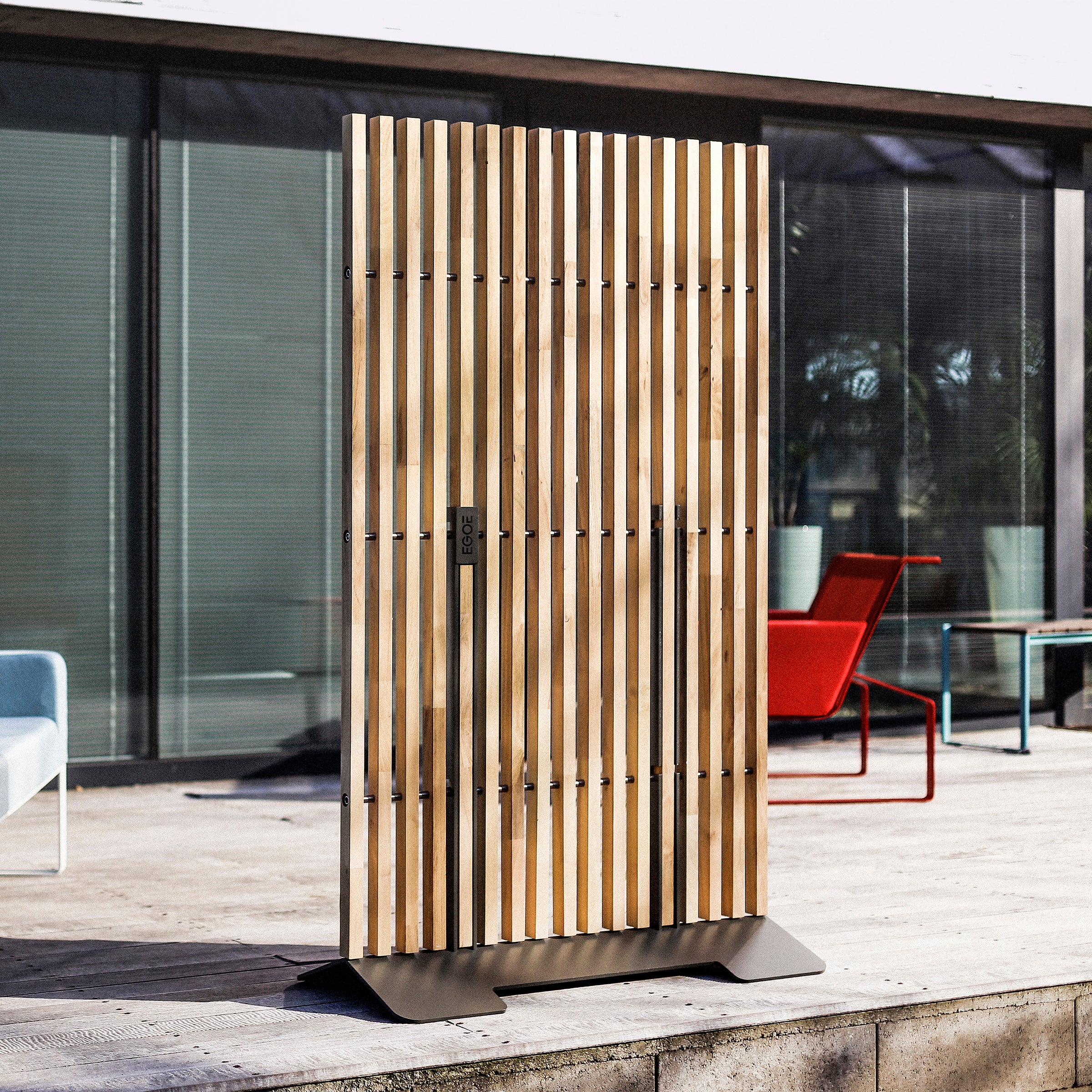 The Mario Screen by Egoe, featuring vertical wooden slats, is set on a concrete patio. In the background, a building with large windows complements the colorful outdoor furniture, including blue and red chairs.