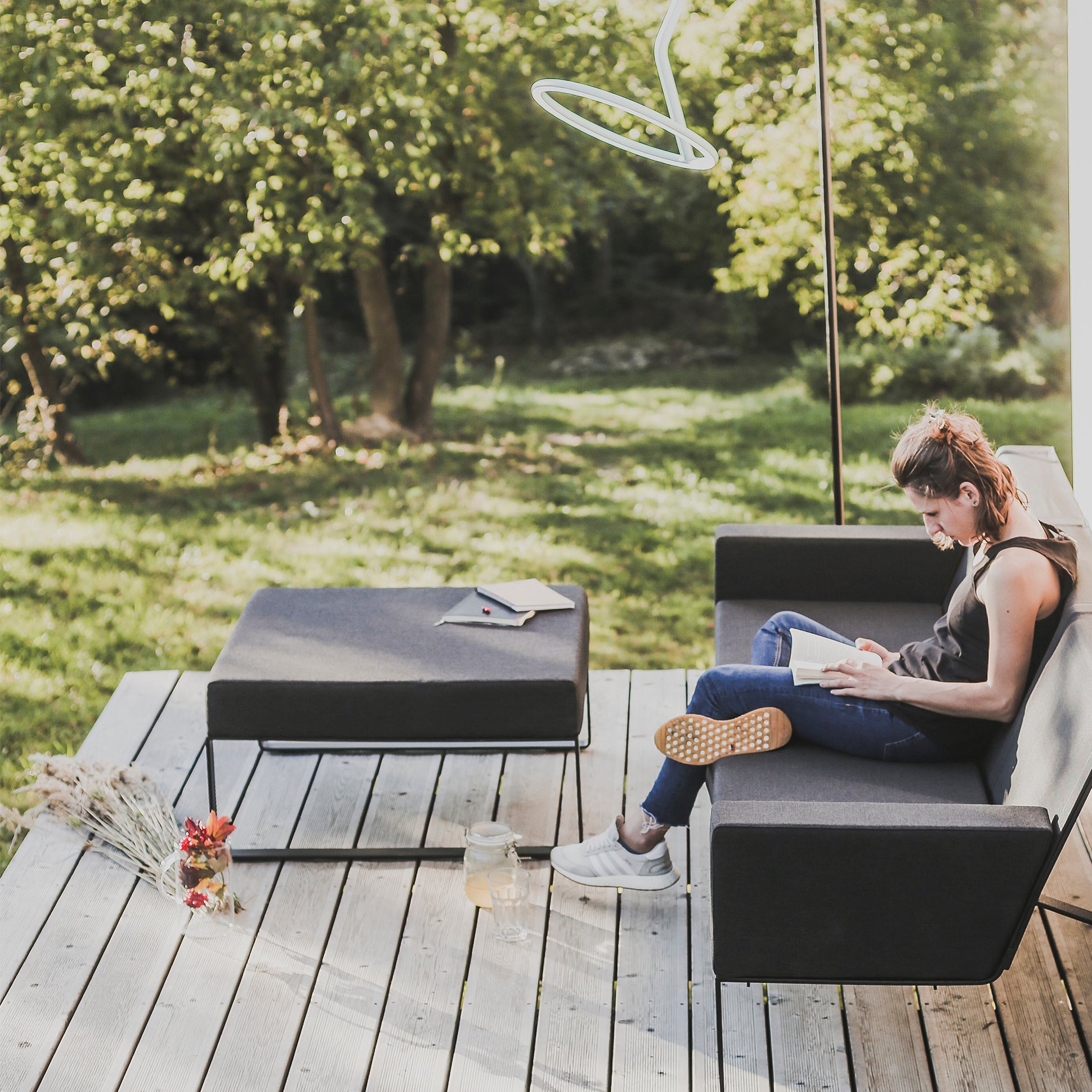 A person relaxes on an Egoe Moja Ottoman on a wooden deck, reading a book. Nearby, a table holds more books and glasses with a jar candle and flowers resting by the galvanised steel frame, all set against lush green trees in the background.