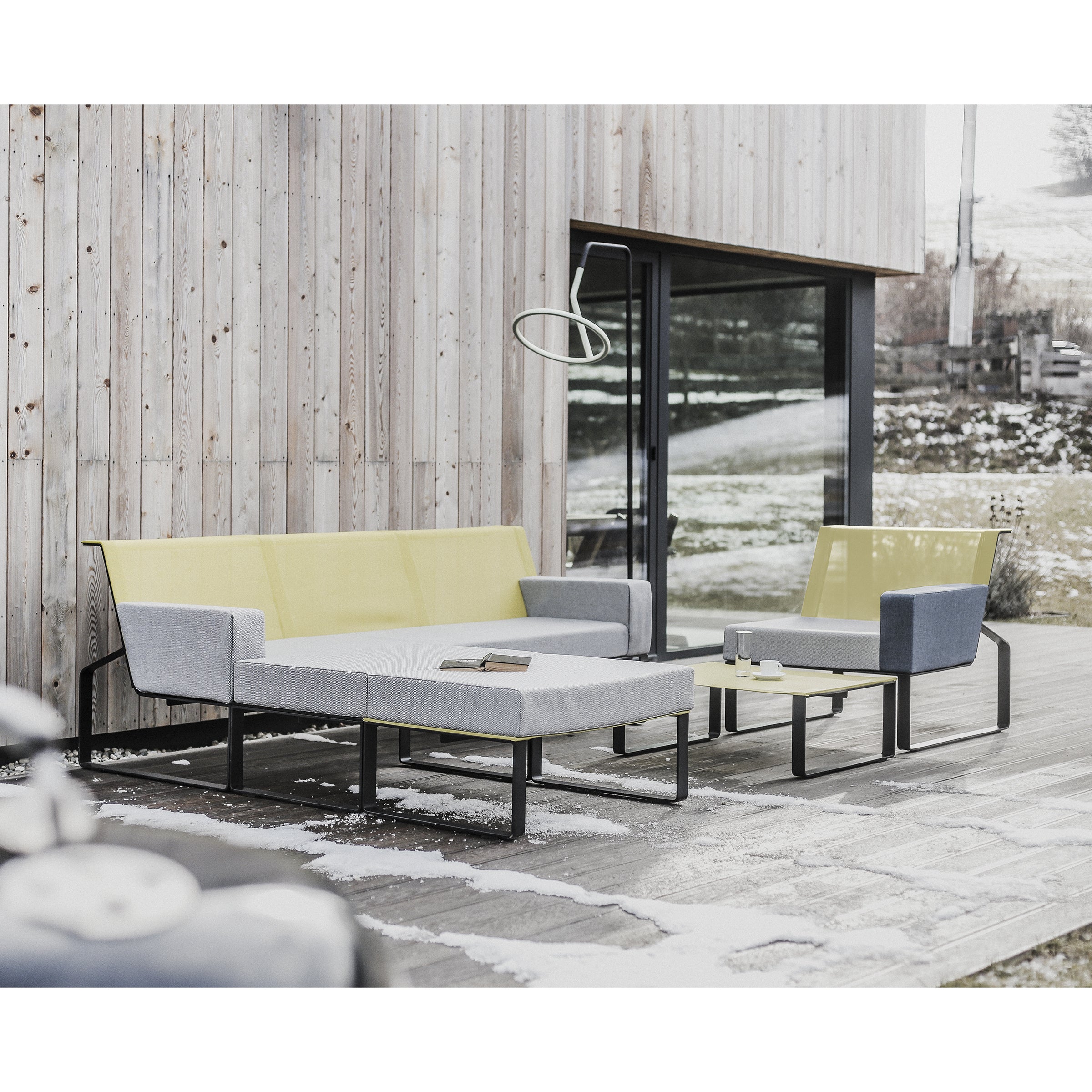 The Egoe Moja Ottoman collection defines the outdoor seating area, showcasing a modern sofa and armchair with gray and yellow cushions on a snow-dusted wooden deck. A galvanized steel table, laden with books, reflects the snowy landscape through a sleek glass facade.