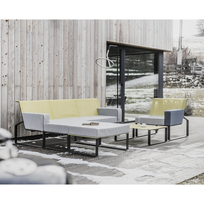 The Egoe Moja Ottoman collection defines the outdoor seating area, showcasing a modern sofa and armchair with gray and yellow cushions on a snow-dusted wooden deck. A galvanized steel table, laden with books, reflects the snowy landscape through a sleek glass facade.