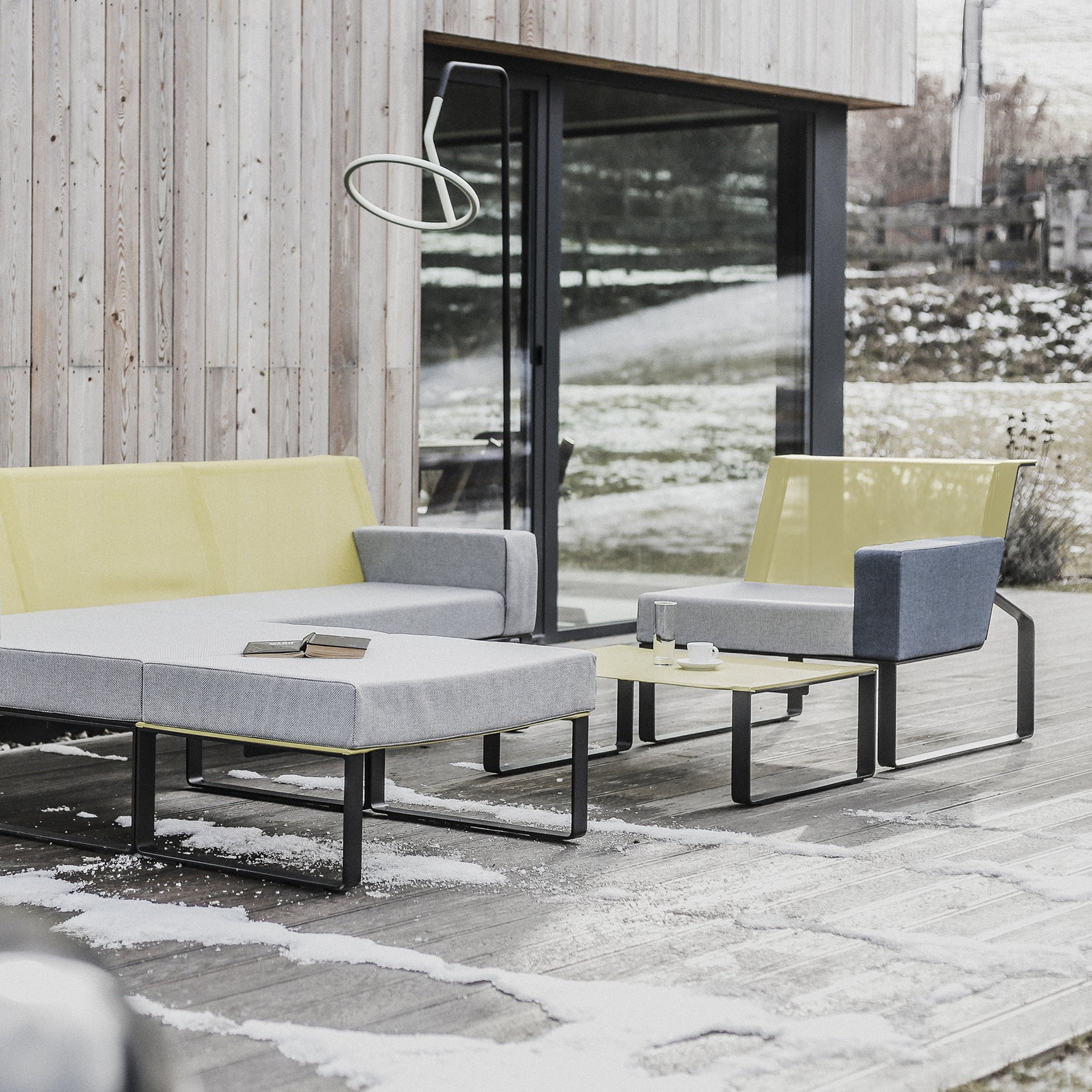 A modern patio features gray and yellow Egoe furniture, including a modular sofa, chair, and Moja Coffee Table topped with books and cups. It has a wooden wall and glass doors, snow-dusted wooden floors, and a circular lamp overhead.