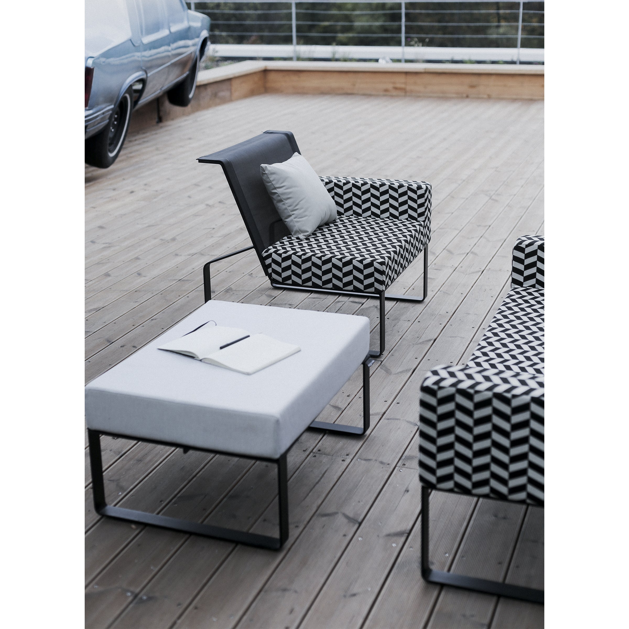 The outdoor seating area on a wooden deck includes a stylish modular collection with a geometric-patterned chair and sofa. Nearby, the Egoe Moja Ottoman holds an open notebook and pen. A classic car adds charm as the railing offers views of the blurred natural landscape.