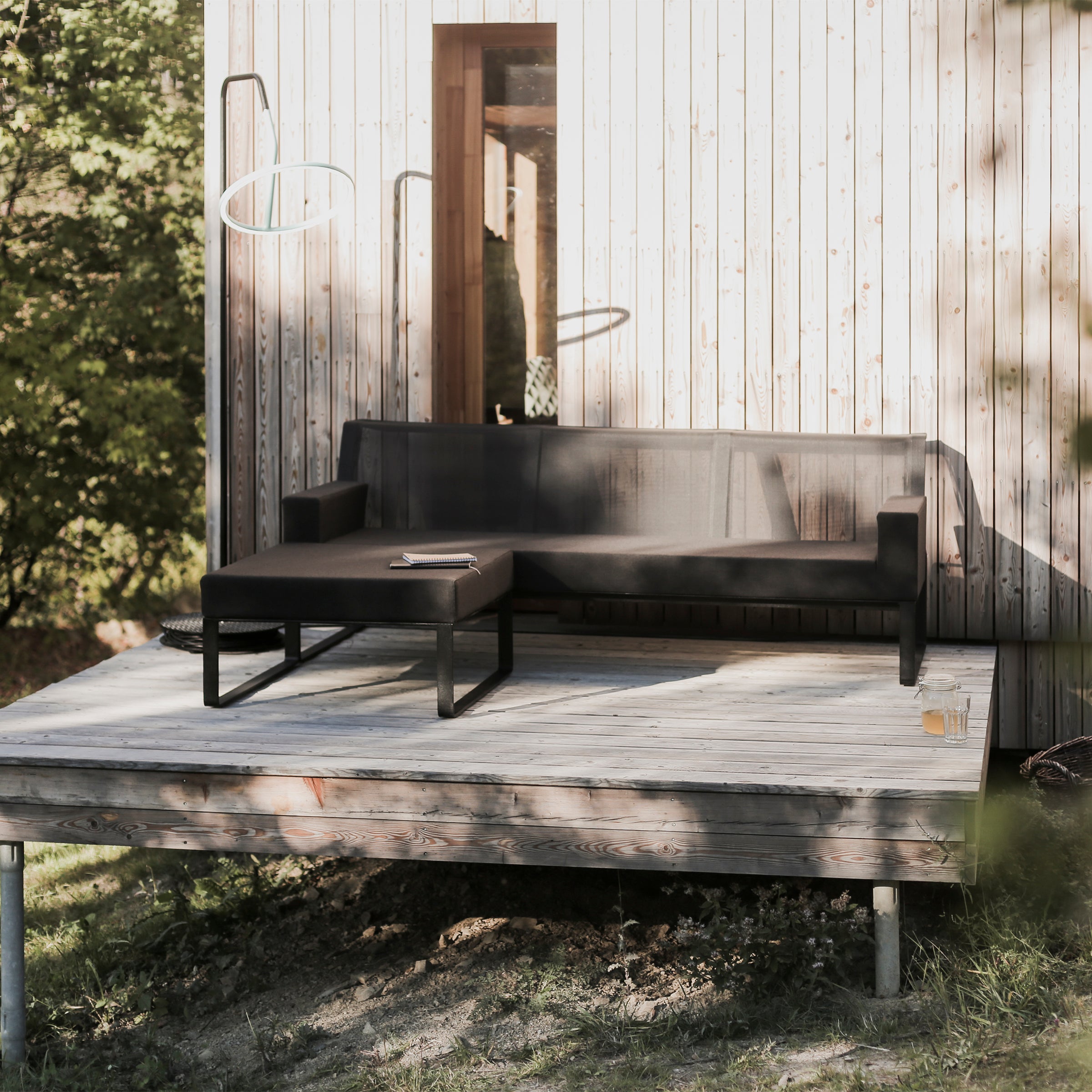 A modern outdoor seating area features a black sofa and the Moja Ottoman by Egoe from a modular collection. The wooden deck is elevated on galvanized steel supports, next to a light wood-paneled building, surrounded by greenery and bathed in gentle sunlight.