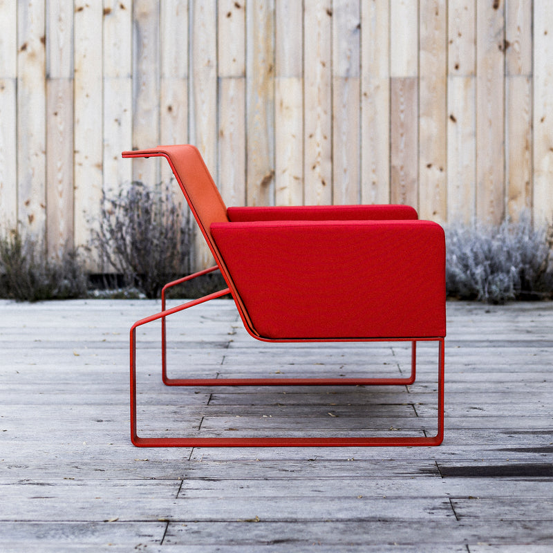 Side view of the Moja Lounge Chair by Egoe, featuring a modern red design with a minimalist galvanized steel frame. Ideal for outdoor seating on a wooden patio, it stands against a light wooden wall with dried plants, showcasing its sleek, contemporary style.
