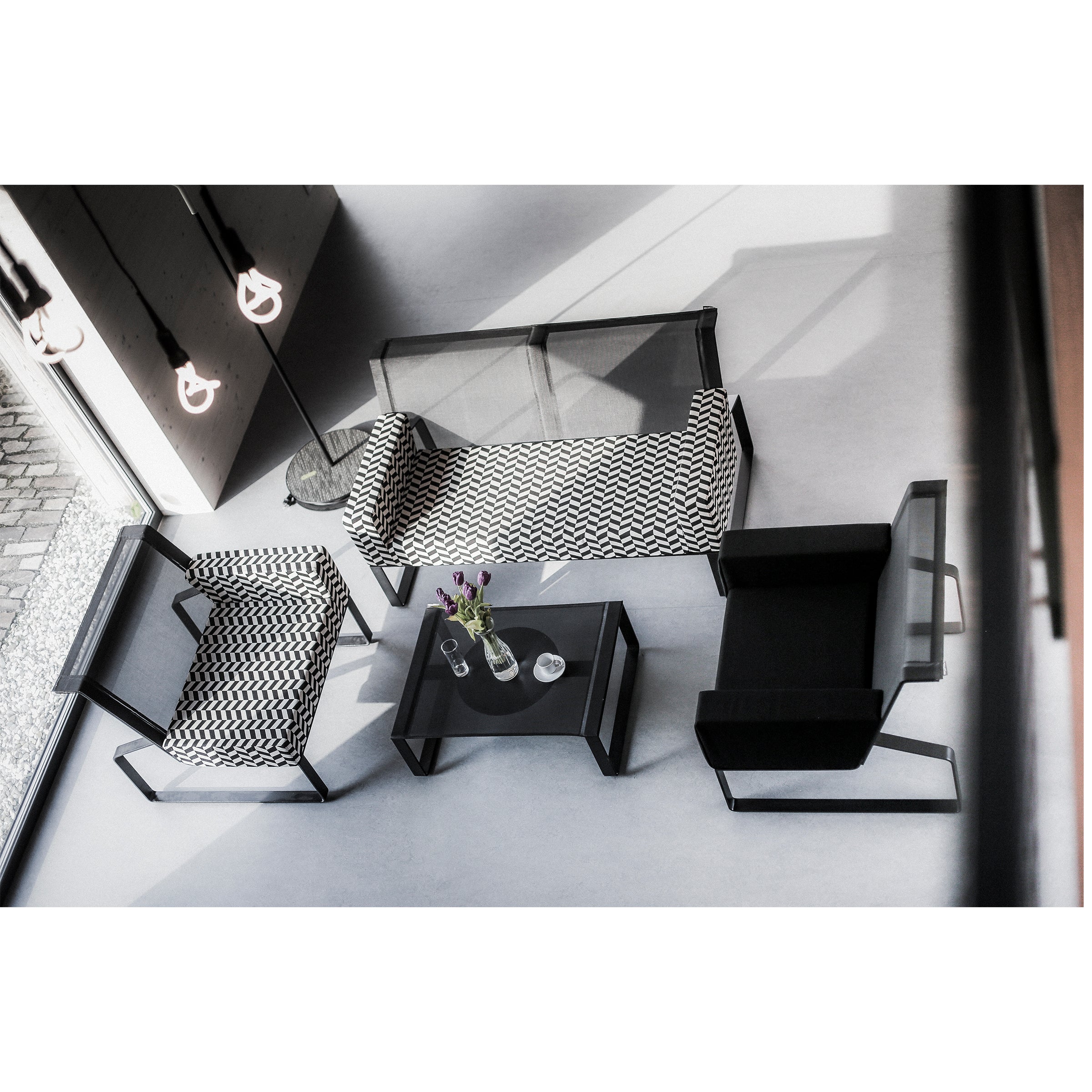 A minimalist modern living room features items from Egoes Moja modular collection: a patterned sofa, two patterned armchairs, a black armchair, and a glass coffee table with flowers. Natural light beautifully illuminates the space.