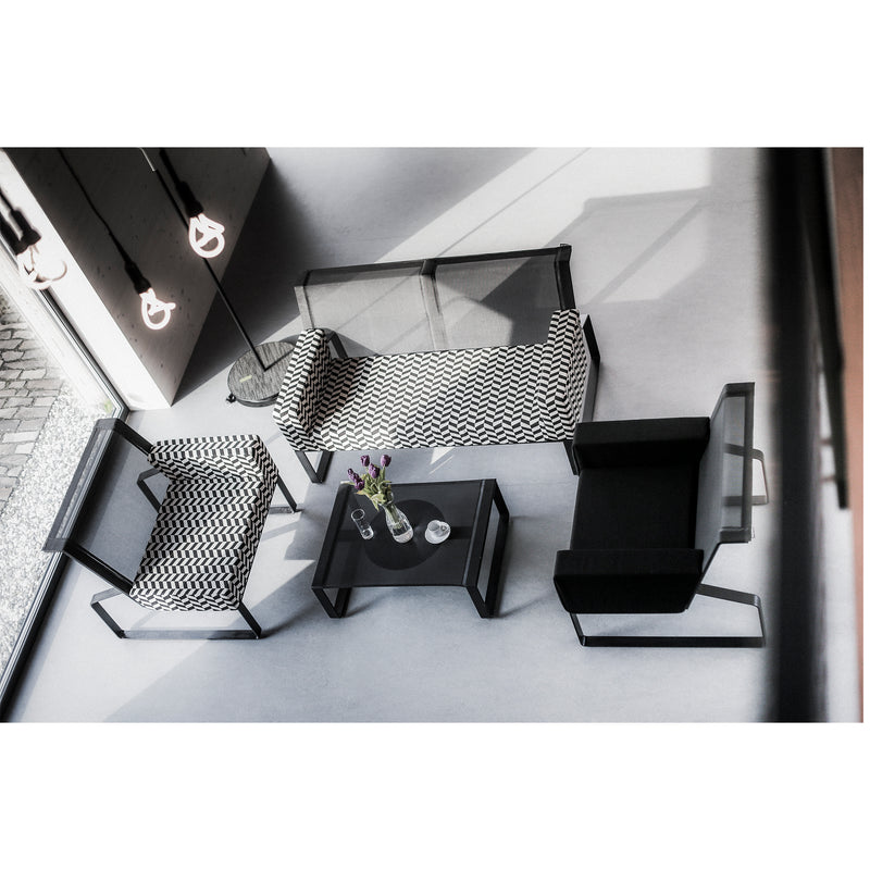 A minimalist modern living room features items from Egoes Moja modular collection: a patterned sofa, two patterned armchairs, a black armchair, and a glass coffee table with flowers. Natural light beautifully illuminates the space.