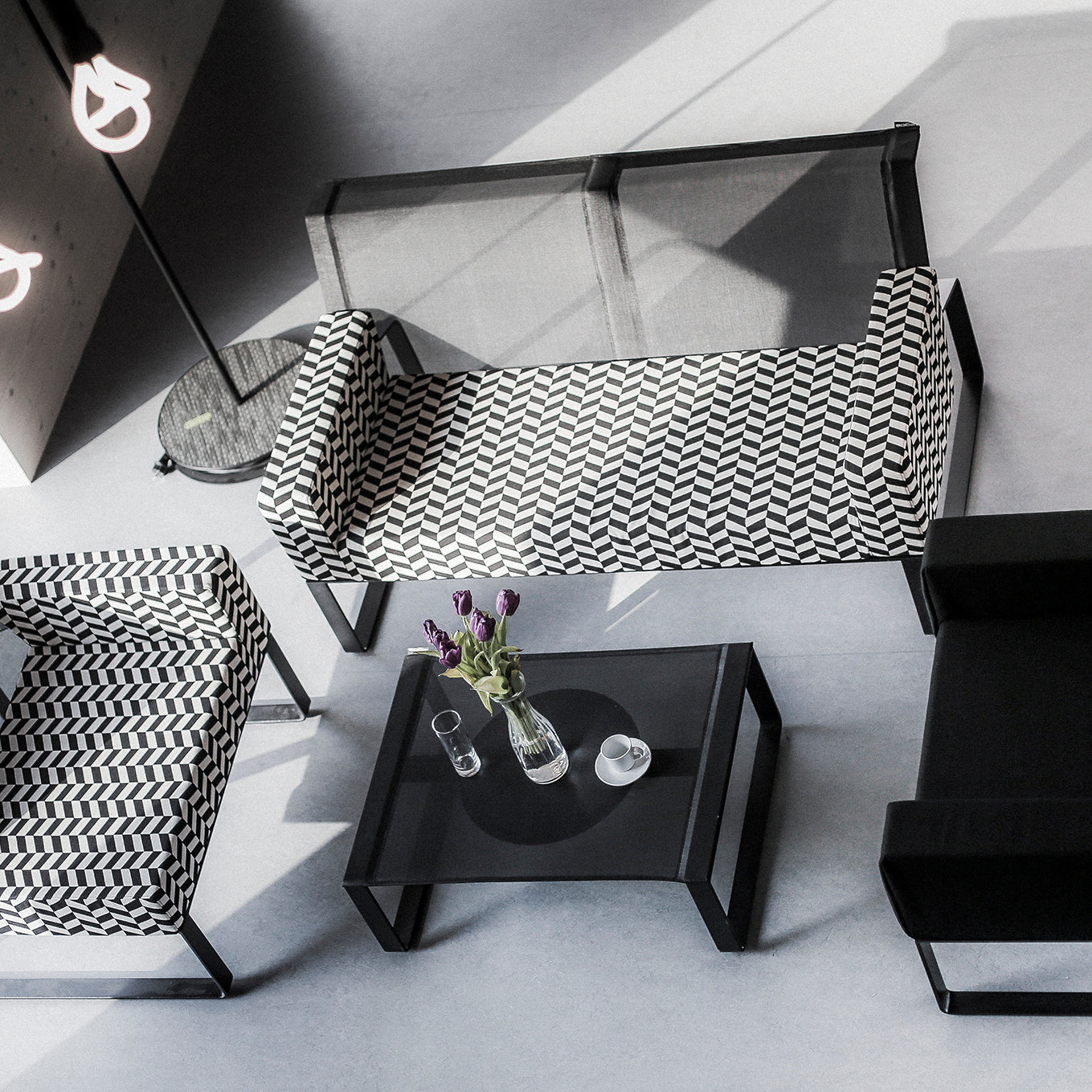 Overhead view of a modern living space featuring an Egoe Moja sofa in black and white pattern, a sleek black sofa, and modular collection elements. A glass-topped coffee table with flowers, pitcher, and cup rests near a floor lamp with looped bulbs on the light-colored floor.